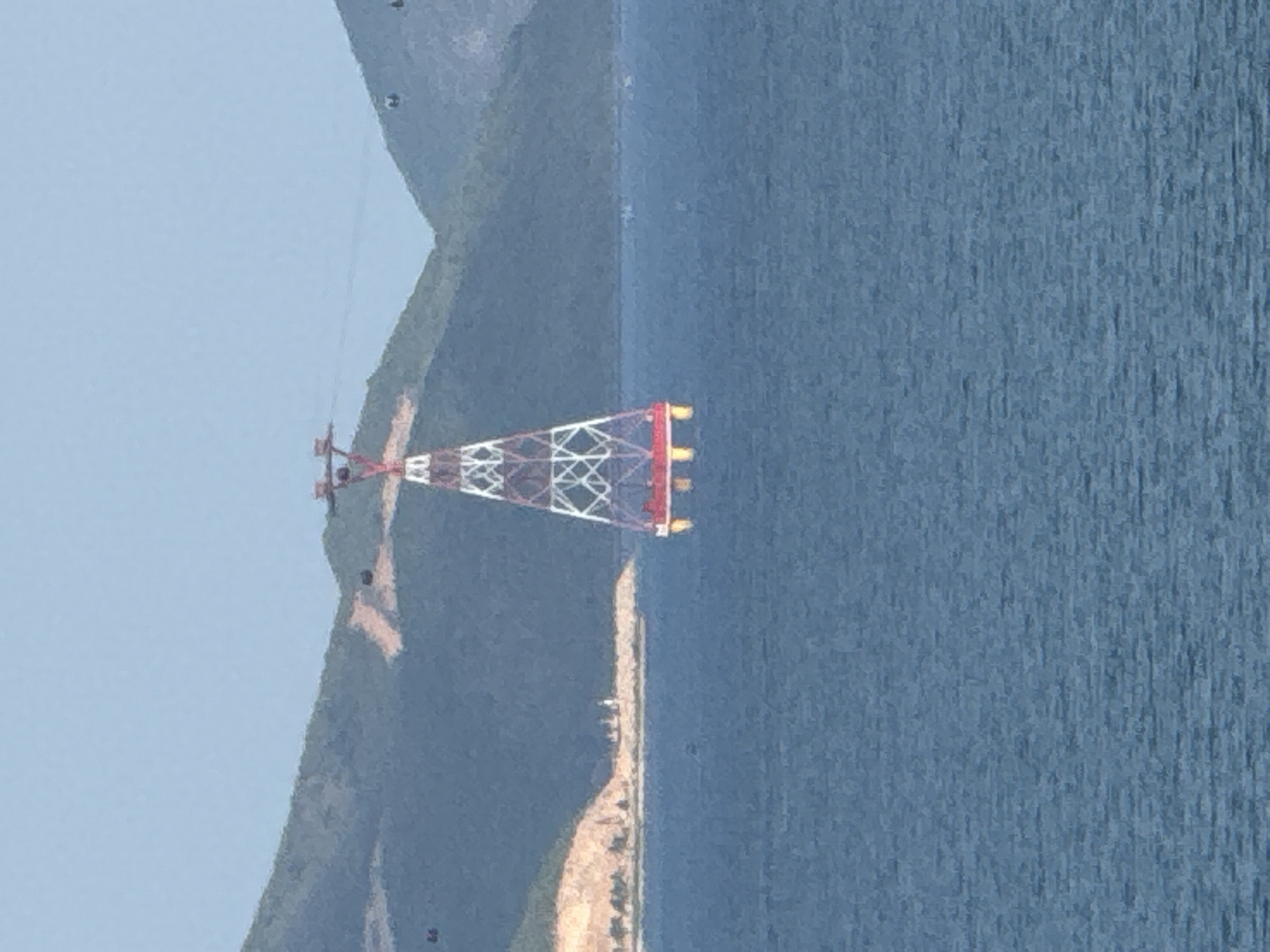 This image shows a cable car tower in the sea off the coast of Nha Trang, Vietnam. The tower is red and white, and it has a cable car system running across it. The tower is supported by four legs that extend into the water. In the background, there are green hills and a blue sky. The water is calm, and there are a few small boats in the distance. The scene appears to be a scenic tourist attraction.