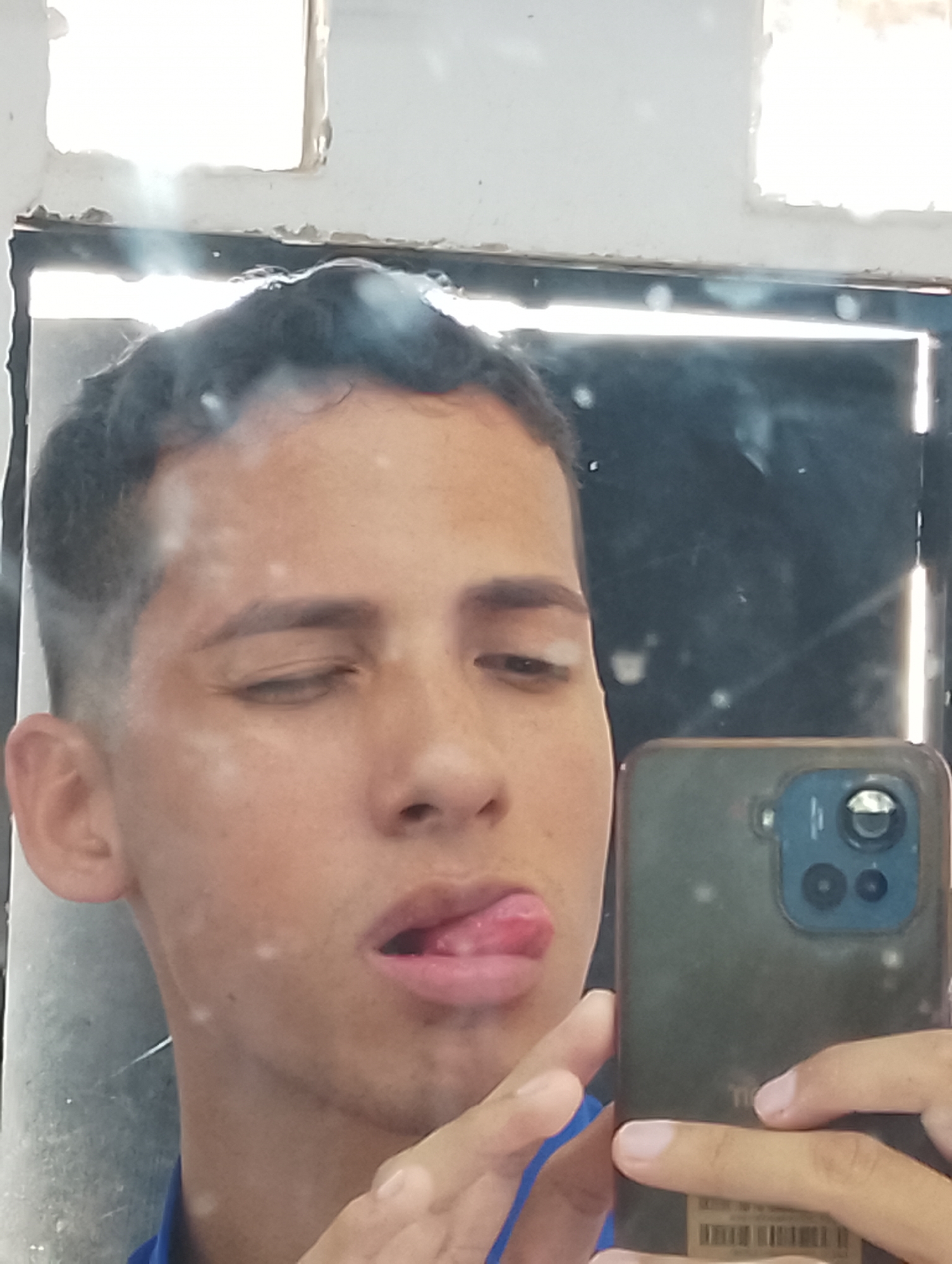 A person is taking a selfie in a mirror. The person has short, dark hair and is wearing a blue shirt. Their tongue is sticking out. The phone in the person's hand is a dark color with multiple camera lenses. The mirror is framed and appears to be in a bathroom or similar space, as there is a partially visible white wall with rectangular cutouts above the mirror. The specific location in Porlamar, Venezuela cannot be determined from the image.