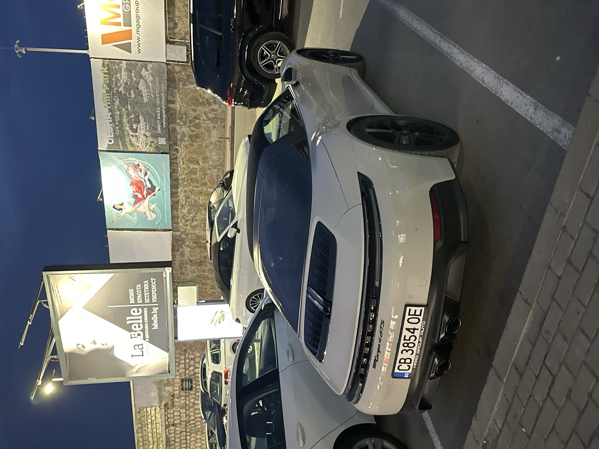 The image shows a parking lot in Varna, Bulgaria, at dusk. Parked vehicles include a white Porsche 911 Targa 4 GTS and a black SUV. Billboards in the background promote "La Belle" aesthetic and dental clinic, "Gesha Hill Park," and "MGA group." The Porsche's license plate reads "CB 3854 OE." The environment is an outdoor parking area with stone and paved surfaces. The "Gesha Hill Park" billboard lists a direct sales number: +359 878236311.