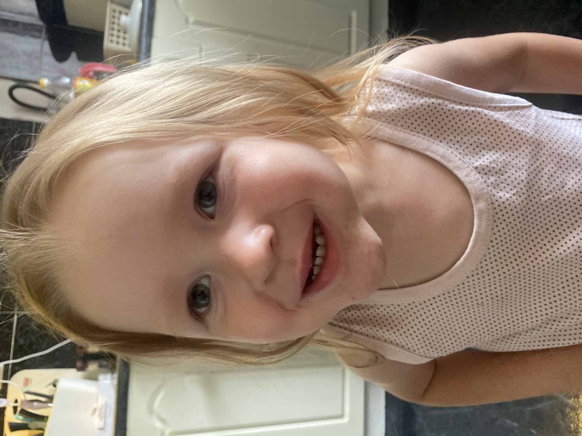 A young Caucasian child with blonde hair and light skin is smiling at the camera. The child is wearing a light pink sleeveless shirt with a pattern of small black dots. The background appears to be a kitchen, with white cabinets and dark countertops visible. There are also various objects on the counter, including what looks like a white appliance and a plastic bottle. The overall scene is brightly lit, suggesting it was taken during the day indoors. There is no specific indication of the location being in Chaikovsky, Russia.