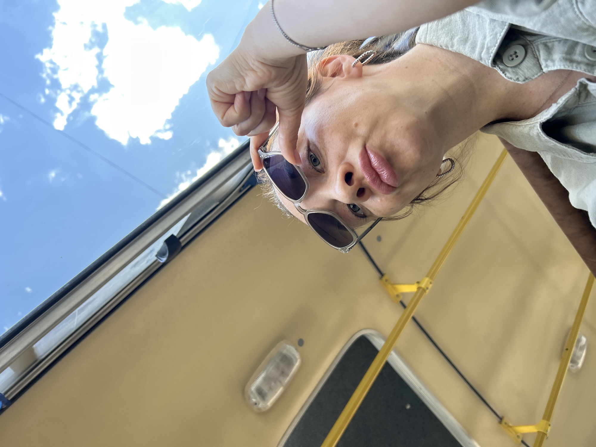 A selfie shows a woman wearing sunglasses and a light-colored button-up shirt, inside a bus in the city of Chaikovsky, Russia. A blue sky with white clouds is visible through the bus window. Yellow handrails and an overhead light are also visible in the interior. The woman is holding her sunglasses.