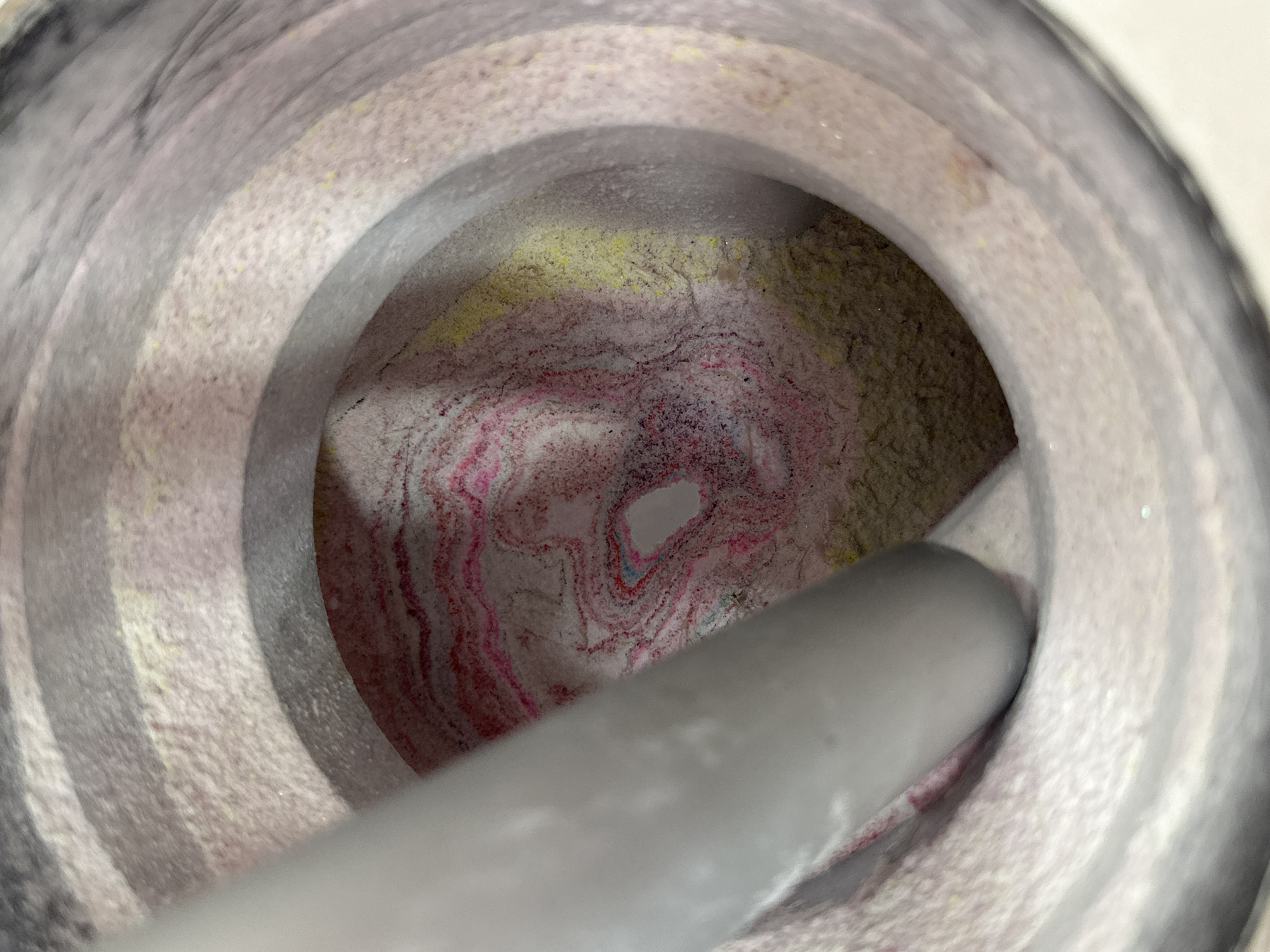 The image shows the interior of a mortar, likely made of stone or ceramic, with a pestle resting inside. The mortar contains a mixture of powdered substances in various colors, including pink, yellow, and brown. The pestle is grey and smooth. The background is out of focus, showing the exterior of the mortar. The location of the scene in Sofia, Bulgaria, is not apparent from the image.