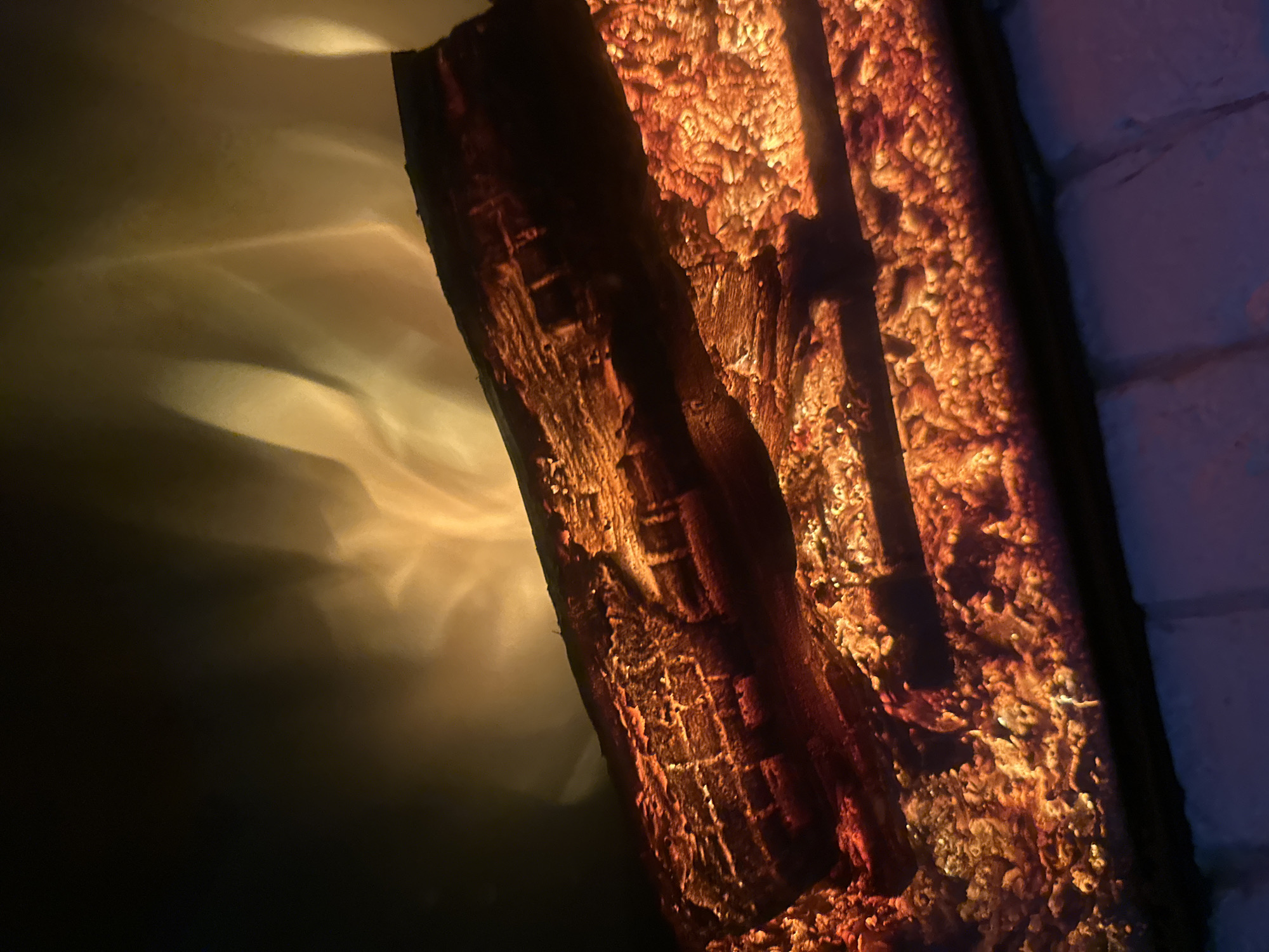 This media file is a close-up, low-angle shot of what appears to be a decorative, artificial log set meant to resemble burning embers. The primary subject is a dark, textured log, possibly made of resin or ceramic, set against a backdrop that suggests a fireplace or hearth. The log itself is detailed with carved lines and imperfections, giving it a realistic appearance.
Illumination is a key element, with warm, orange and yellow light emanating from within and around the log, mimicking the glow of a fire. This light casts dramatic shadows and highlights on the surface of the log and on a textured, possibly brick or stone wall visible on the right side of the frame. The light also creates wavy patterns on a dark, out-of-focus area to the left, suggesting a diffusely lit background or perhaps a reflection.
The overall scene evokes a sense of warmth and coziness, often associated with a fireplace on a cool evening. The lighting creates a moody and inviting atmosphere. There is no visible text in the image. The focus is entirely on the textural qualities of the artificial log and the interplay of light and shadow.