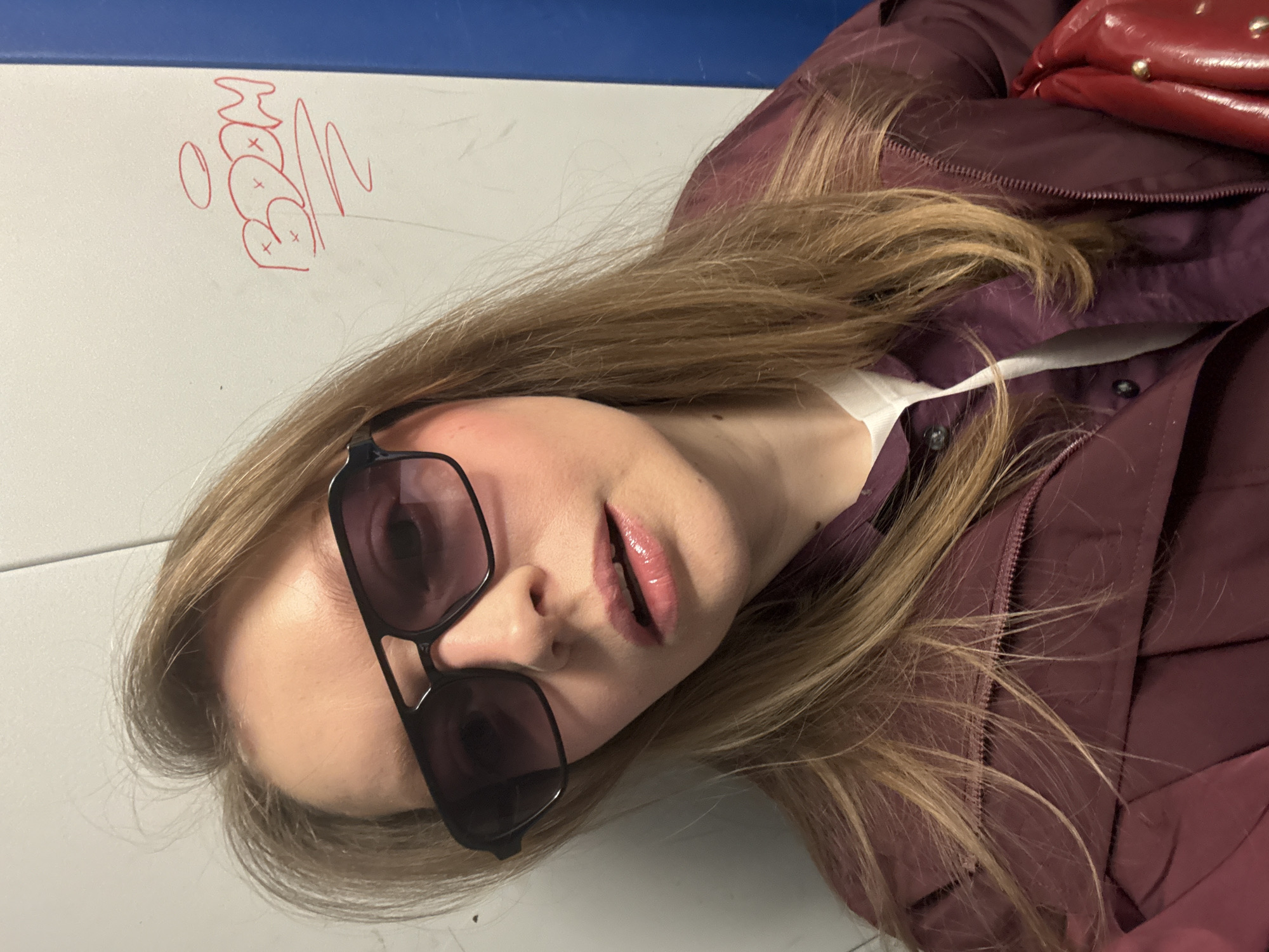 A woman wearing sunglasses and a burgundy jacket lies on a white surface with blue trim, likely a table. A red purse rests on the jacket near her shoulder. Red graffiti-like markings are visible on the table near her head. There is no information about the location being in Izhevsk, Russia.