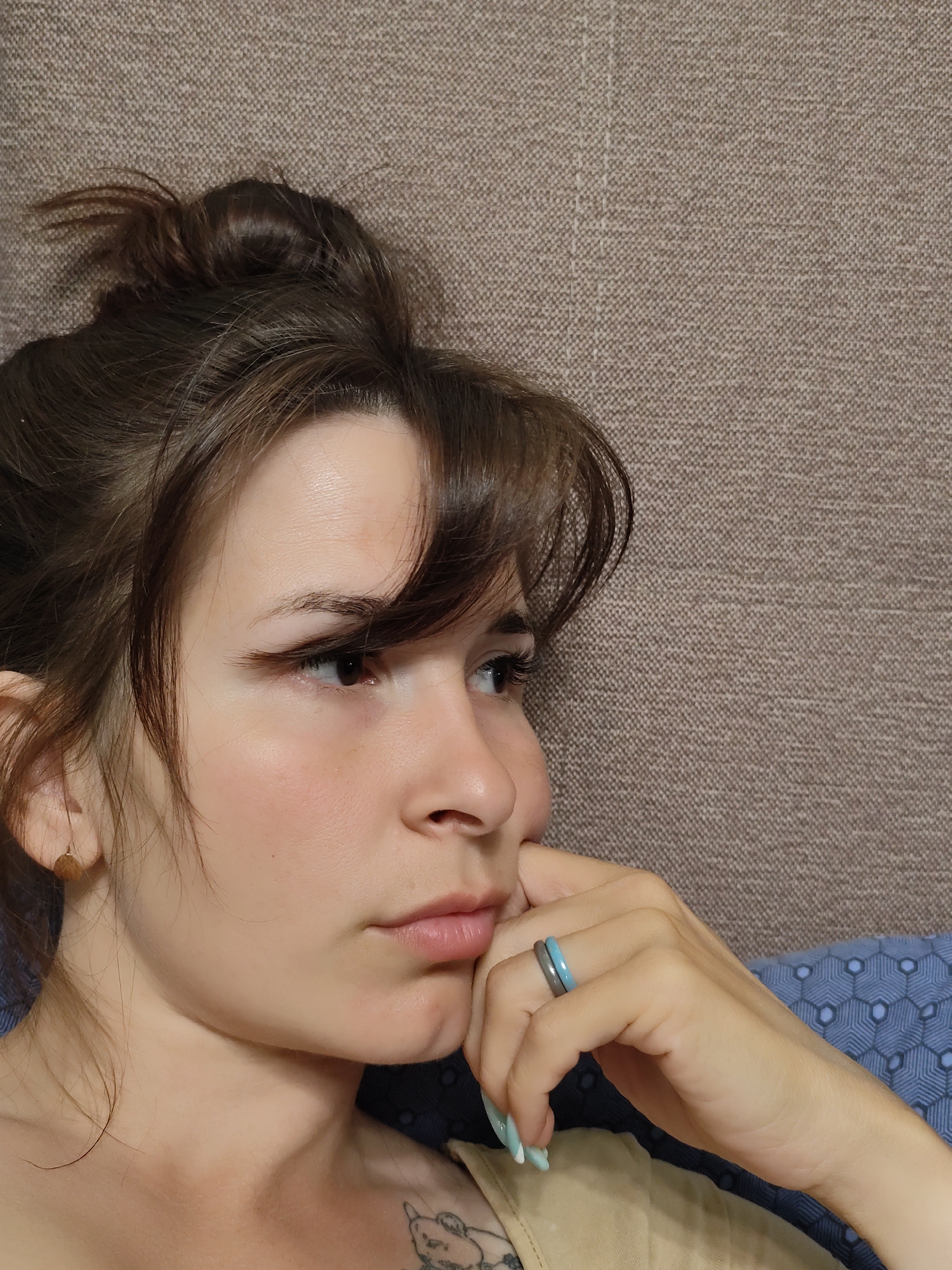 A close-up shot shows a woman with brown hair styled in a messy bun, gazing thoughtfully to the right. She rests her chin on her hand, which is adorned with two rings. The background consists of a neutral-toned fabric behind her head and a blue fabric with a geometric pattern near her shoulder. A tattoo is visible on her upper chest.
The location is not discernible from the image.