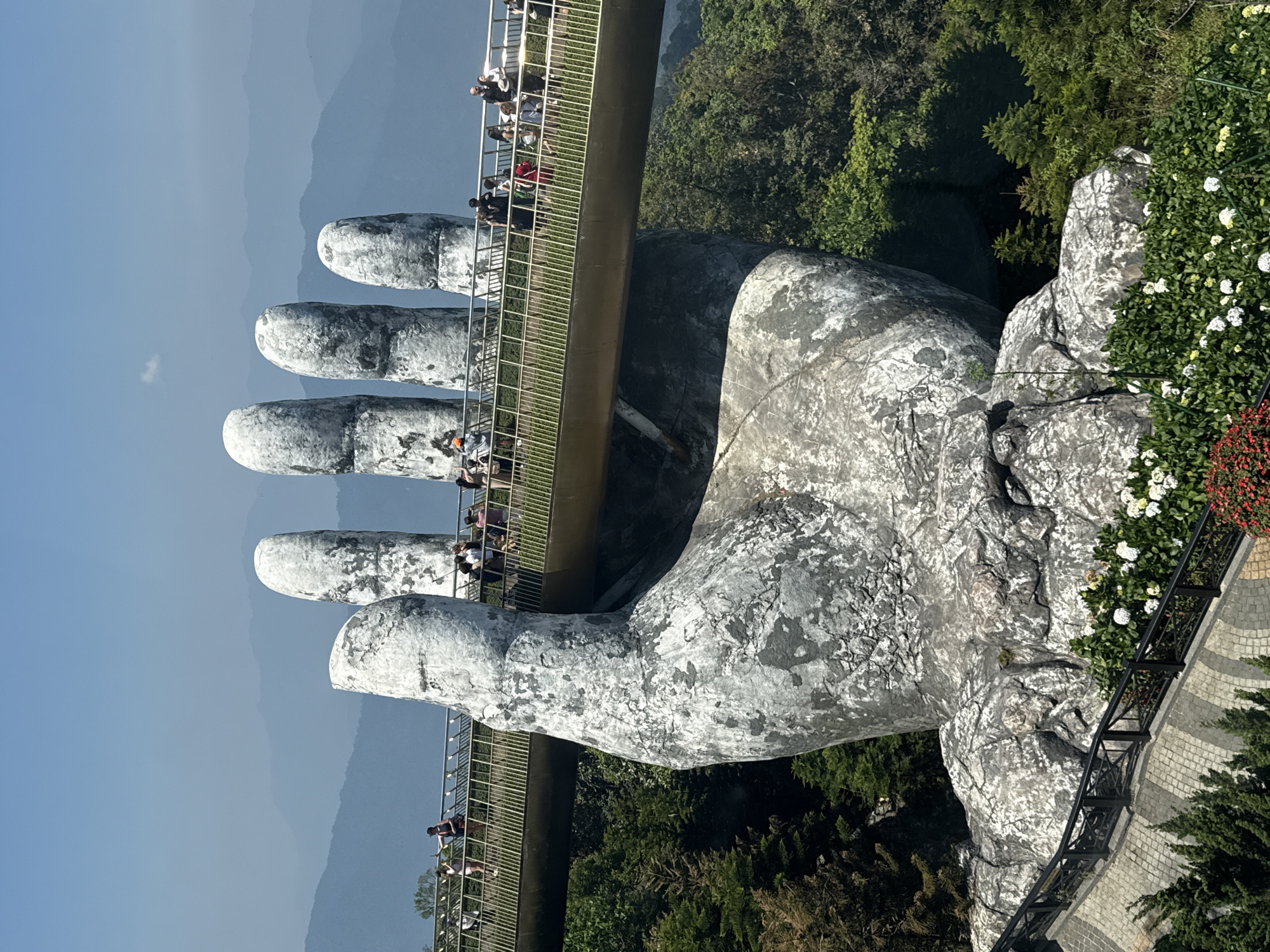 The image depicts the Golden Bridge (Cầu Vàng) at the Bà Nà Hills resort in Đà Nẵng, Vietnam. The bridge is held up by two giant, sculpted stone hands. Numerous people are seen walking along the bridge. The surrounding environment includes dense foliage and distant mountains under a clear sky. The bridge's design incorporates metal railings. The sculpture's surface shows textured details, with a weathered appearance. Flowers and foliage are planted alongside the paved walkway that approaches the sculpture.