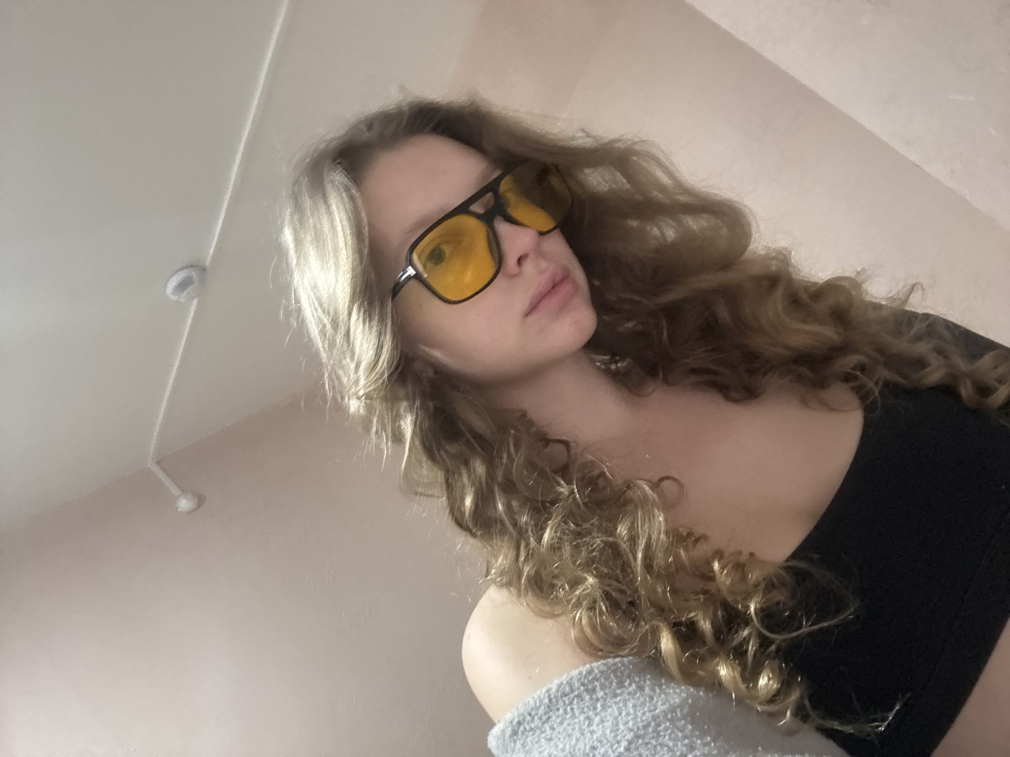 A young woman with curly blonde hair is taking a selfie in a room. She is wearing large, black-framed sunglasses with yellow lenses, and a black top, partially covered by a grey knit sweater. The background is a light-colored wall, and a white ceiling with a smoke detector is visible in the upper left corner. The image appears to have been taken indoors, possibly in a residential setting. There are no specific visual cues to identify the location as Izhevsk, Russia.