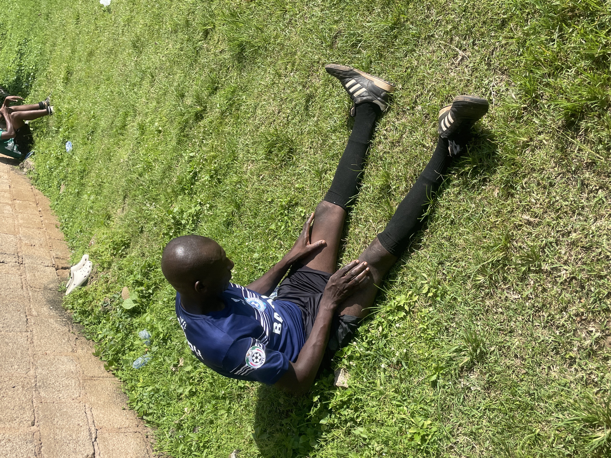 A Black man is sitting on a grassy field with his legs extended in front of him, his body leaning slightly forward and his head bowed. He is wearing a blue and white soccer jersey, black shorts, black socks, and black soccer cleats with white stripes. The jersey has a logo with a globe and soccer ball on the left sleeve. The man appears to be stretching or resting. The environment is a sunny outdoor scene with a stone wall in the background to the left. There are several plastic water bottles and a white shoe near the wall. The grass is green and appears to be well-maintained, suggesting an outdoor sports facility or park in Jos, Nigeria.