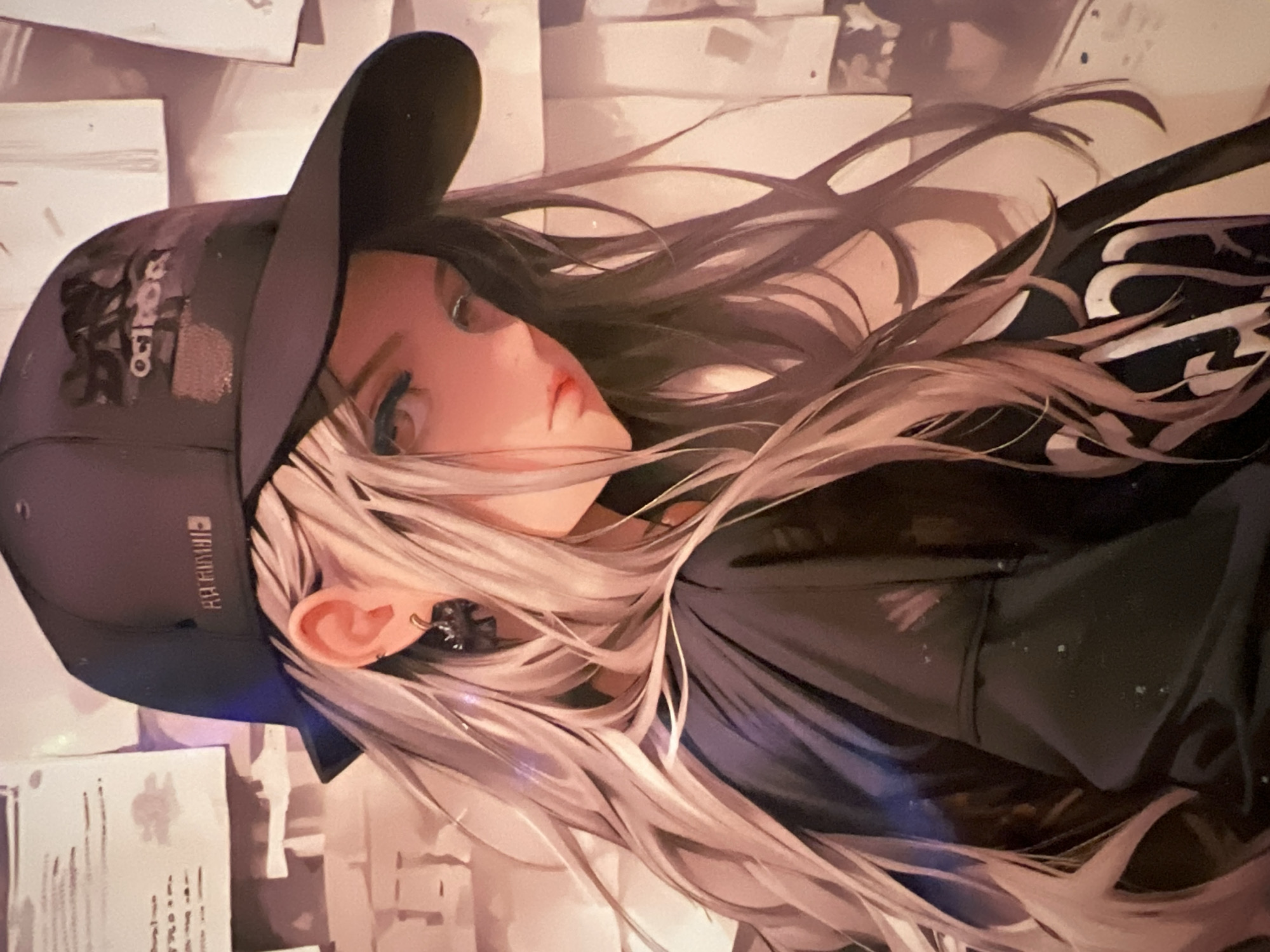 This media file is an artistic rendering of a young woman with long, flowing, light-colored hair. She is wearing a dark baseball cap with white text on the front that appears to be a logo. She also has a small earring in her visible ear. The woman's face is partially turned away from the viewer, but her eyes are visible, and she has a neutral expression. Her hair is styled in loose waves, and some strands are falling across her face and shoulders. She is wearing a dark garment, possibly a jacket or shirt. The background is indistinct, appearing as a collage of abstract shapes and lines, suggesting a busy or urban environment. There is some text visible on the side of the baseball cap, which is in a rectangular box. The overall impression is one of a stylized portrait, possibly for a fashion or artistic context. The provided location context of Minsk, Belarus, does not offer specific visual cues within the image itself.