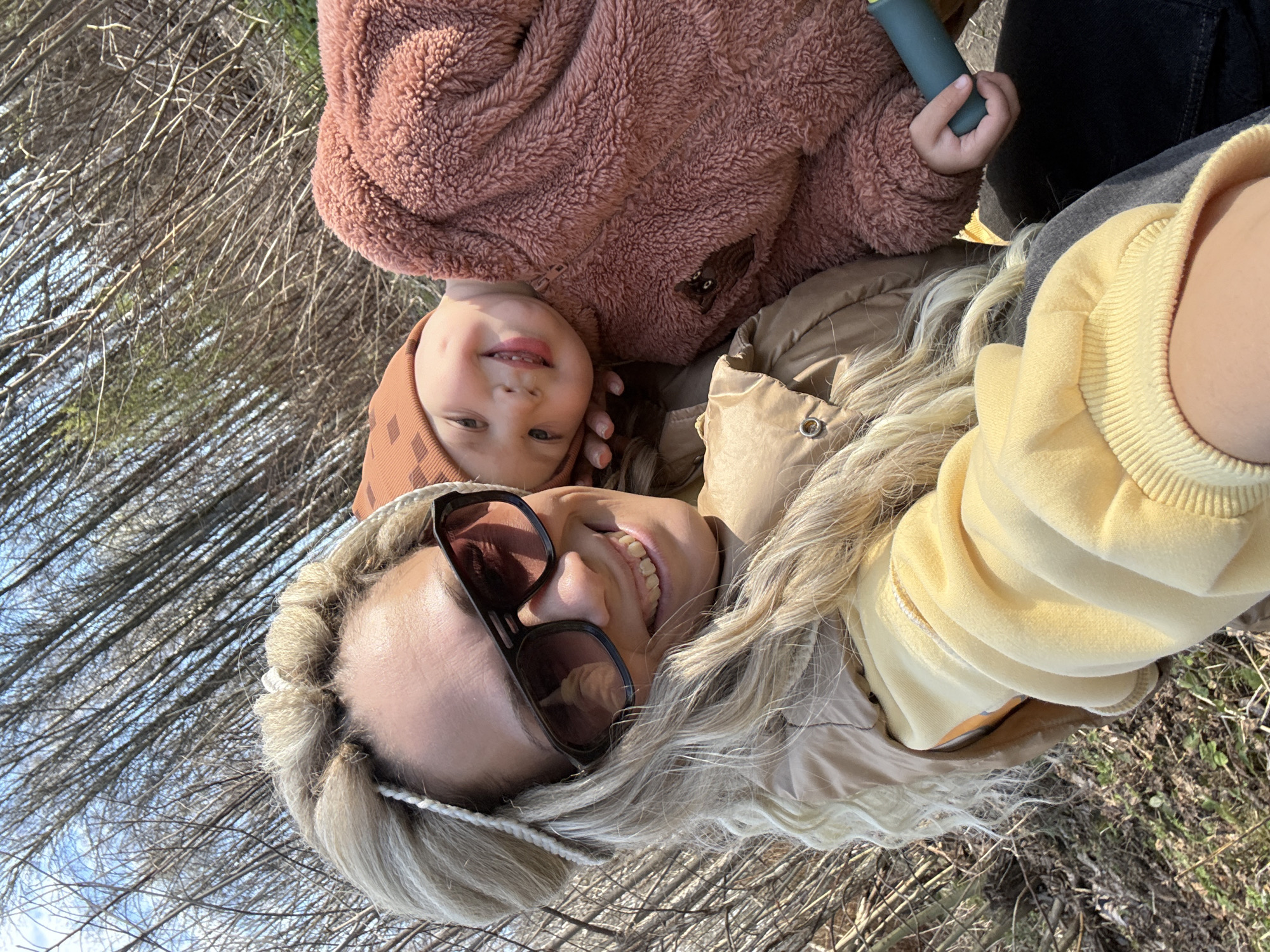 The image shows a fair-skinned woman with long blonde hair, wearing sunglasses and a yellow hooded jacket, lying on her back outdoors with a young child. The child, who also has fair skin, wears a brown hooded outfit and a fluffy pink jacket. The background appears to be a wooded area with bare trees. They are likely situated on the ground. The exact location in Rusanovo, Russia cannot be determined from the image. The interaction seems amicable, possibly a selfie taken by the woman with the child.