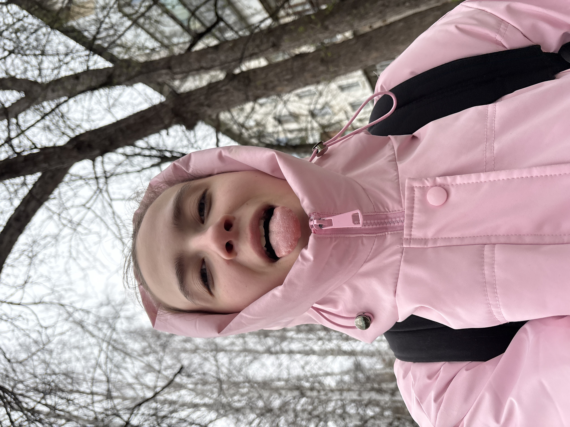 A close-up, low-angle shot captures a young person outdoors in Chaikovsky, Russia. The individual, likely a female, is wearing a bright pink hooded jacket with the hood up, and black backpack straps are visible over their shoulders. They are looking towards the camera with a playful expression, sticking their tongue out. The tongue appears to have some texture or coating. The jacket features a pink zipper, a drawstring with a silver toggle on the hood, and a pink snap button on the lower part.
The background consists of a wintry or early spring landscape. Bare tree branches, characteristic of deciduous trees in colder seasons, are prominent against a bright, overcast sky. Some blurred architectural elements, possibly buildings, can be faintly discerned through the branches in the mid-ground. The overall lighting suggests it is daytime. The image is oriented vertically with the person's head tilted, indicating a dynamic or impromptu selfie-style shot. There is no visible text in the image.