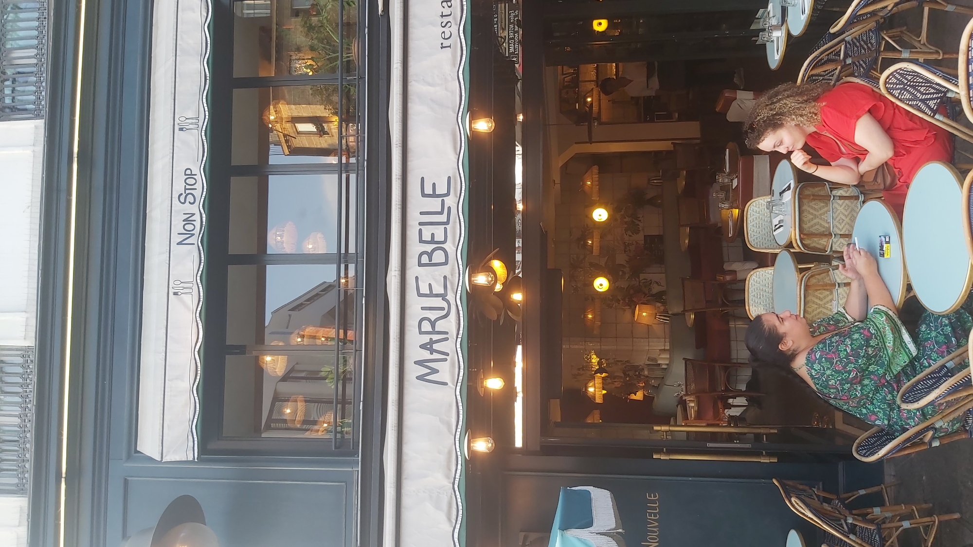 The image shows a street-level view of a Parisian restaurant named "MARIE BELLE" on a sunny day. The restaurant has a large awning with the name in bold, blue lettering. Beneath the awning, the entrance to the restaurant is visible, with a warm glow emanating from within. Several tables with chairs are set up outside the restaurant, some occupied by patrons. Two women are seated at a table in the foreground, engaged in conversation. One woman, wearing a green, floral-patterned dress, holds a cigarette. The other woman, in a red dress, rests her chin on her hand. The street appears to be a cobblestone or paved surface, and there are other cafe tables and chairs visible in the background, suggesting a bustling street scene. The architecture of the building suggests it's in a historic district of Paris, possibly near a well-known landmark given the style of the building and streetscape.