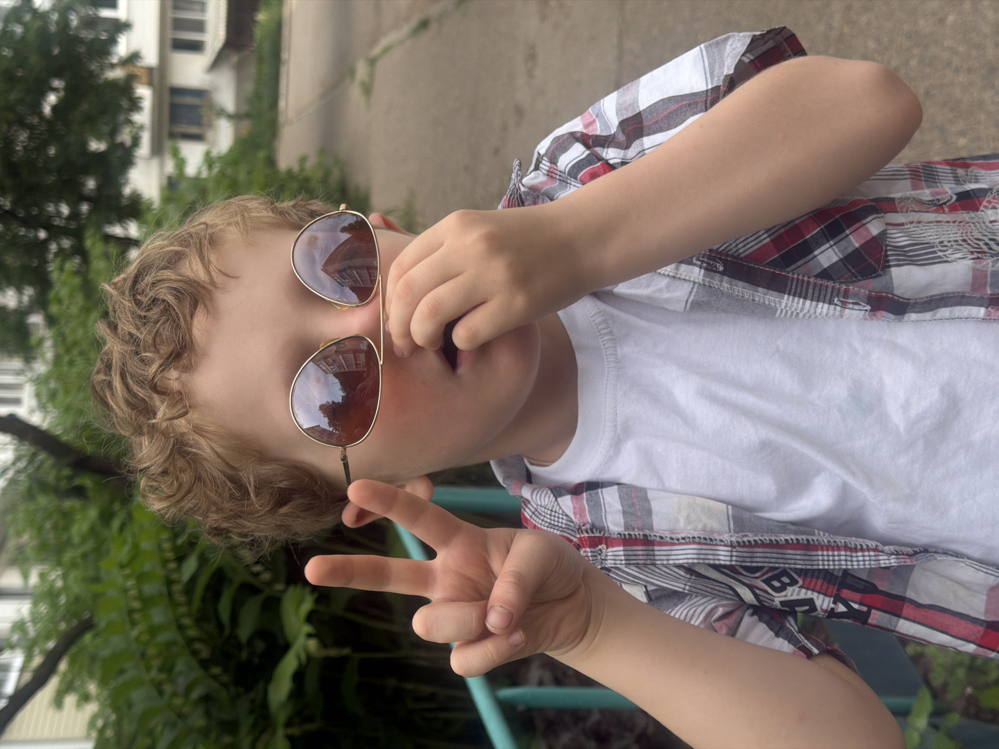 A young child is outdoors, holding sunglasses up to their face, and giving a peace sign with their left hand. They are wearing a white t-shirt under an open, plaid, button-down shirt. The reflection in their sunglasses shows a multi-story building. The environment includes greenery, a sidewalk or paved area, and a building in the background. The location is Chaikovsky, Russia.