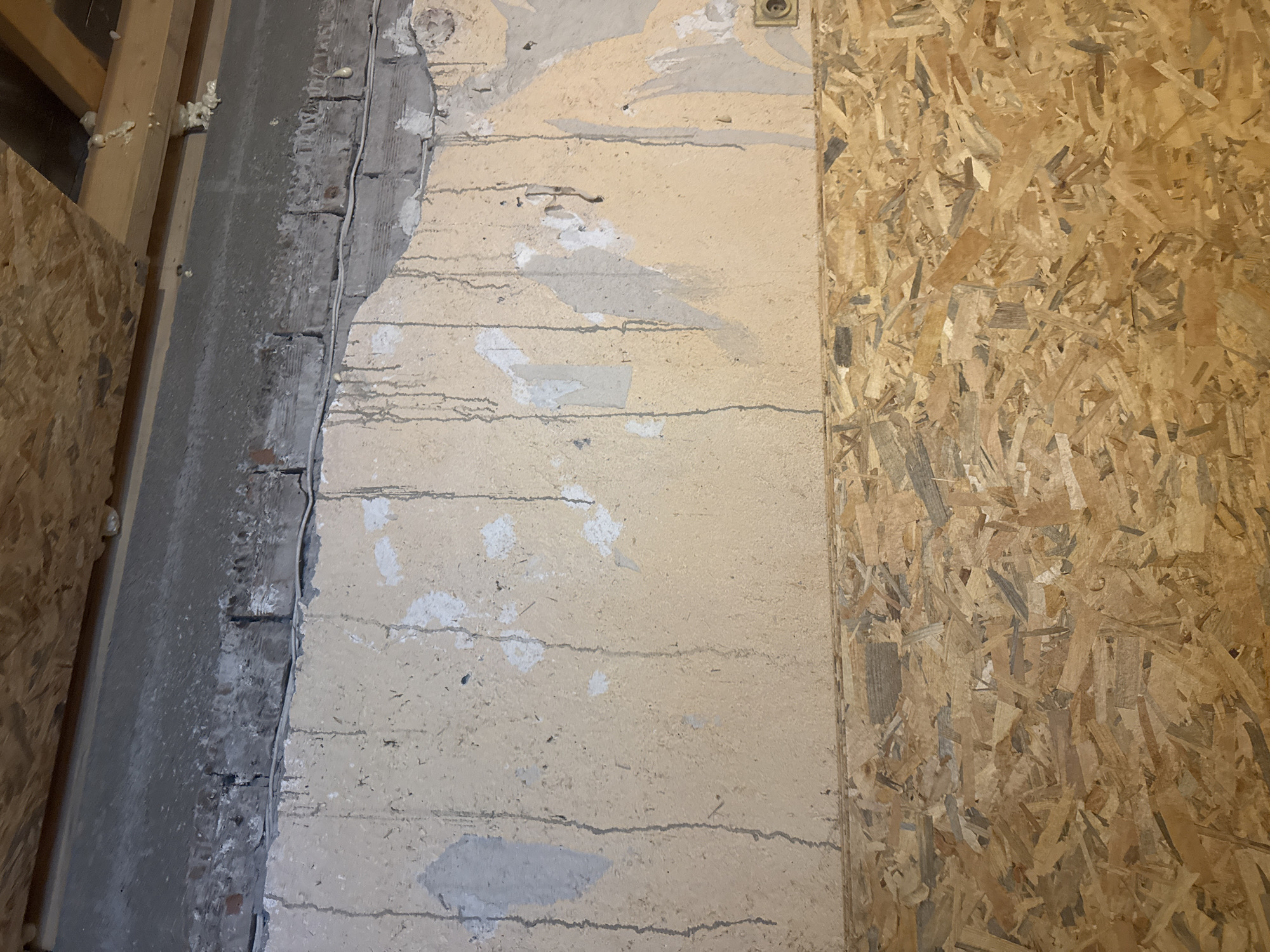 The image displays an interior wall and ceiling, reportedly located in Sarnevo, Bulgaria, depicting an unfinished or deteriorated state.
The uppermost section features an exposed ceiling constructed from oriented strand board (OSB) panels, supported by horizontal wooden beams. Irregular deposits of yellowish-white spray foam insulation are visible adhering to these wooden beams and the OSB, particularly towards the right side.
Immediately below the ceiling structure, a horizontal band of rough, dark grey material, consistent with concrete or unpainted plaster, traverses the upper wall. Beneath this grey band, a course of exposed and weathered brickwork, partially obscured by lighter grey remnants, is visible. A thin, white electrical wire runs horizontally across the wall, positioned just below the exposed brickwork, and appears to be secured in places.
The main wall surface beneath the brickwork is covered in a light beige or tan textured plaster or paint. This surface exhibits extensive damage, with numerous irregular patches where the top layer has peeled or fallen away, revealing a lighter grey substrate or underlying masonry. Distinct dark grey or black vertical streaks, consistent with dried water runoff or staining, are prominent across the beige wall surface, originating from the upper wall and brickwork and extending downwards. Towards the lower right of this section, a circular, brass-colored electrical outlet or junction box is partially visible.
The lower portion of the wall is uniformly clad with oriented strand board (OSB) panels, displaying the characteristic variegated pattern of compressed wood flakes in shades of light to medium brown. This OSB cladding appears relatively intact and newer than the wall section above it.
No individuals or direct actions are present in the scene. The overall environment suggests an interior space undergoing construction, renovation, or experiencing significant disrepair, marked by visible raw materials and evidence of water damage.