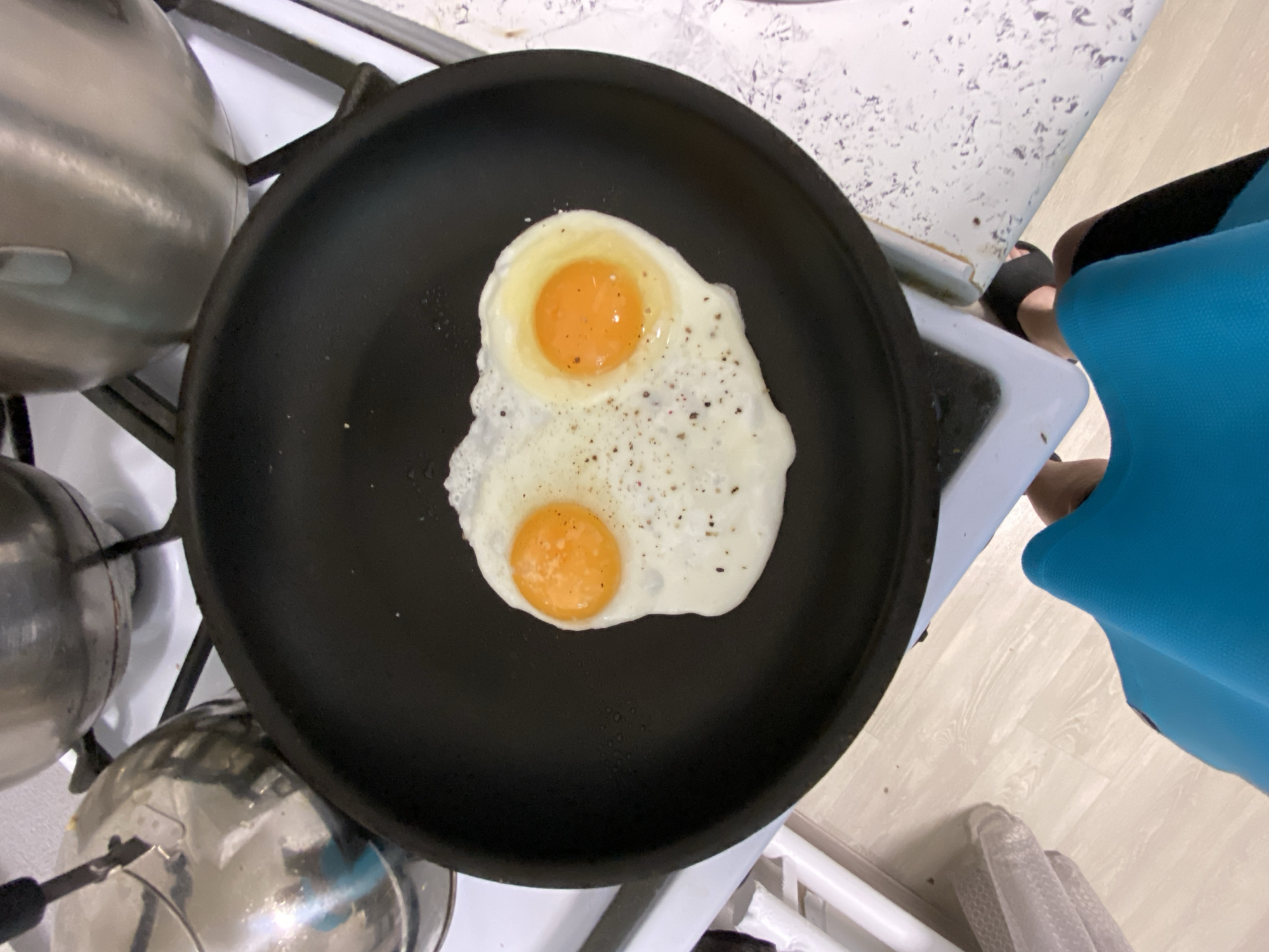 A top-down view shows a black frying pan on a white stovetop, containing two fried eggs with yolks still runny. The eggs are seasoned with pepper. To the left are metal pots and pans. A person wearing black sandals and a blue dress stands to the right of the stove. The location is a kitchen, presumably indoors. There is no definitive information to confirm the location is Noviy, Russia.