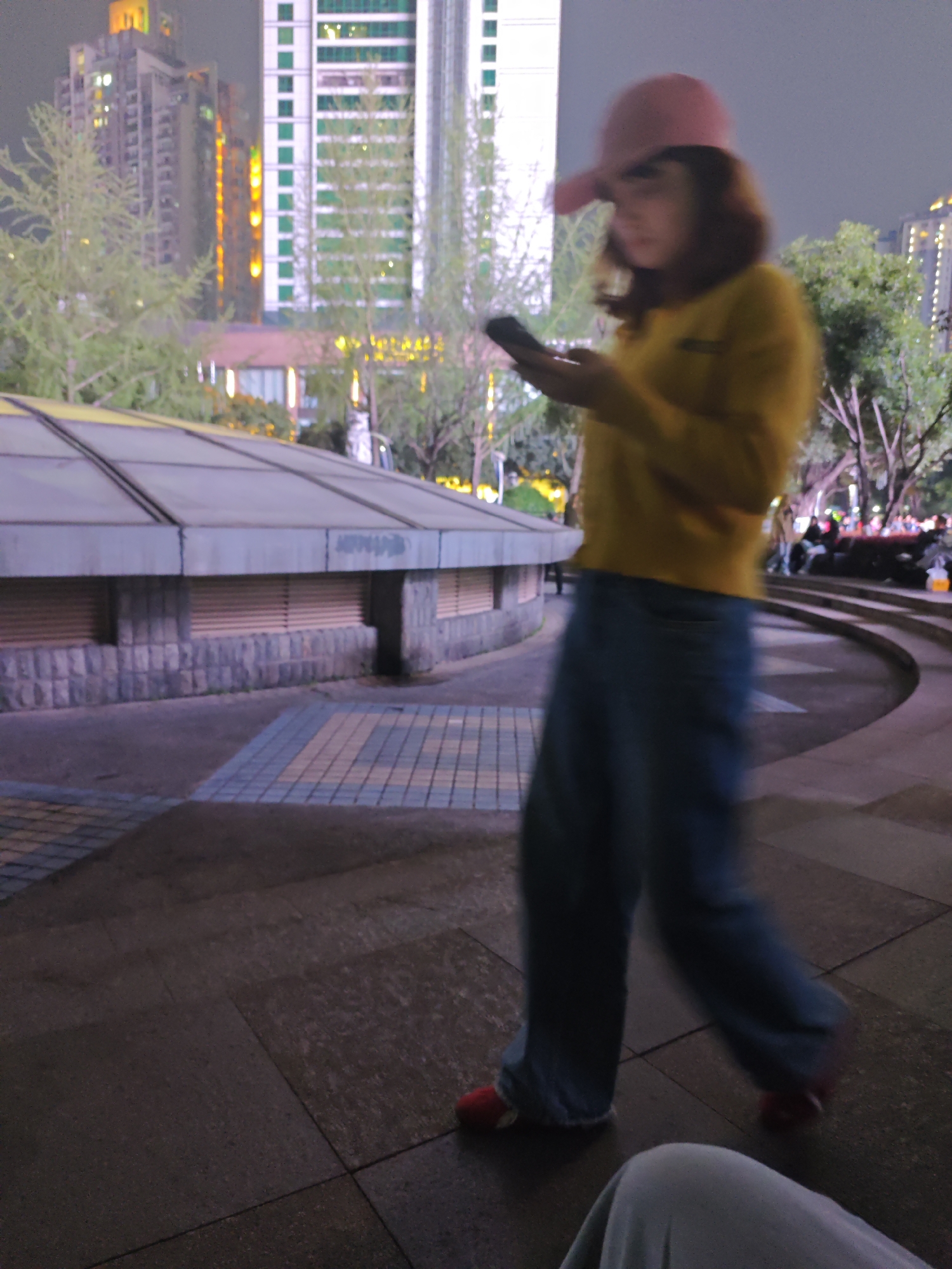 A blurred figure of a person wearing a pink baseball cap, yellow long-sleeved shirt, and blue jeans is walking and looking at a cell phone in Liangjiang, China. The person is walking in a park, on dark paving stones, near a low building with a slanted roof made of multiple rectangular sections. The building's base is made of gray brick. In the background are tall buildings with illuminated windows. Trees are scattered throughout the park. Other people are sitting on steps in the background. A person's knee is visible in the bottom center of the image. The scene is at night.