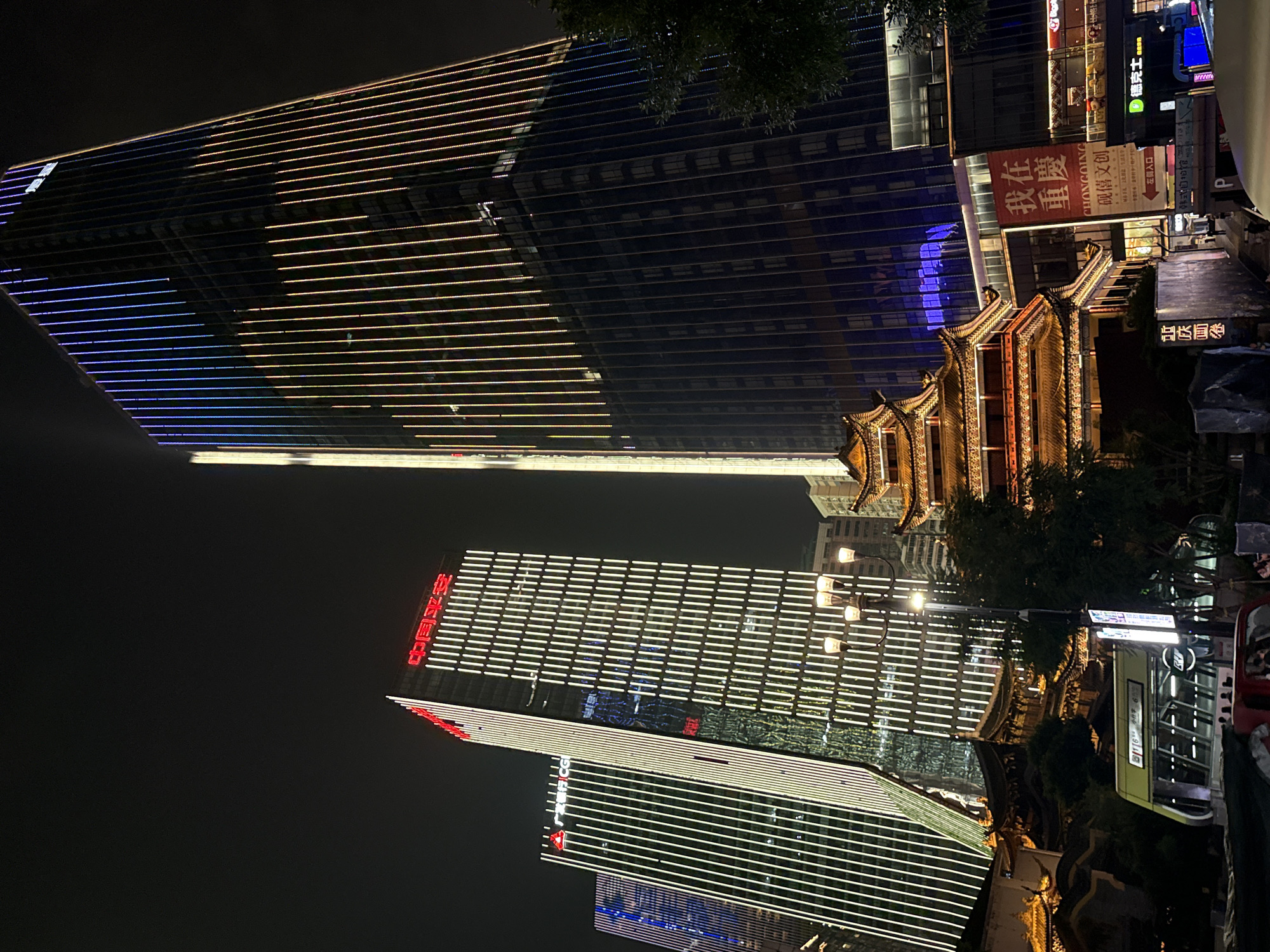 The image captures a nighttime urban scene in Yuzhong District, China, dominated by illuminated skyscrapers and traditional architecture. A large, modern skyscraper on the right is adorned with vertical LED lights in yellow, orange, and blue, creating a vibrant spectacle against the dark sky. To its left, two other skyscrapers also feature prominent vertical lighting in white and green. In the foreground, a multi-tiered, traditional Chinese building with glowing golden accents stands out, its ornate roofline contrasting with the sharp angles of the modern structures. A street lamp with multiple globes illuminates the area in front of the traditional building. Below the skyscrapers and traditional architecture, a street-level view reveals a building entrance with Chinese characters, a bus, and what appears to be a subway entrance with a sign displaying Chinese text and a circular logo. The overall atmosphere is one of urban dynamism, blending historical and contemporary elements under the night sky. There are no visible people engaged in specific actions, but the presence of a bus and subway entrance suggests public transit and pedestrian activity.