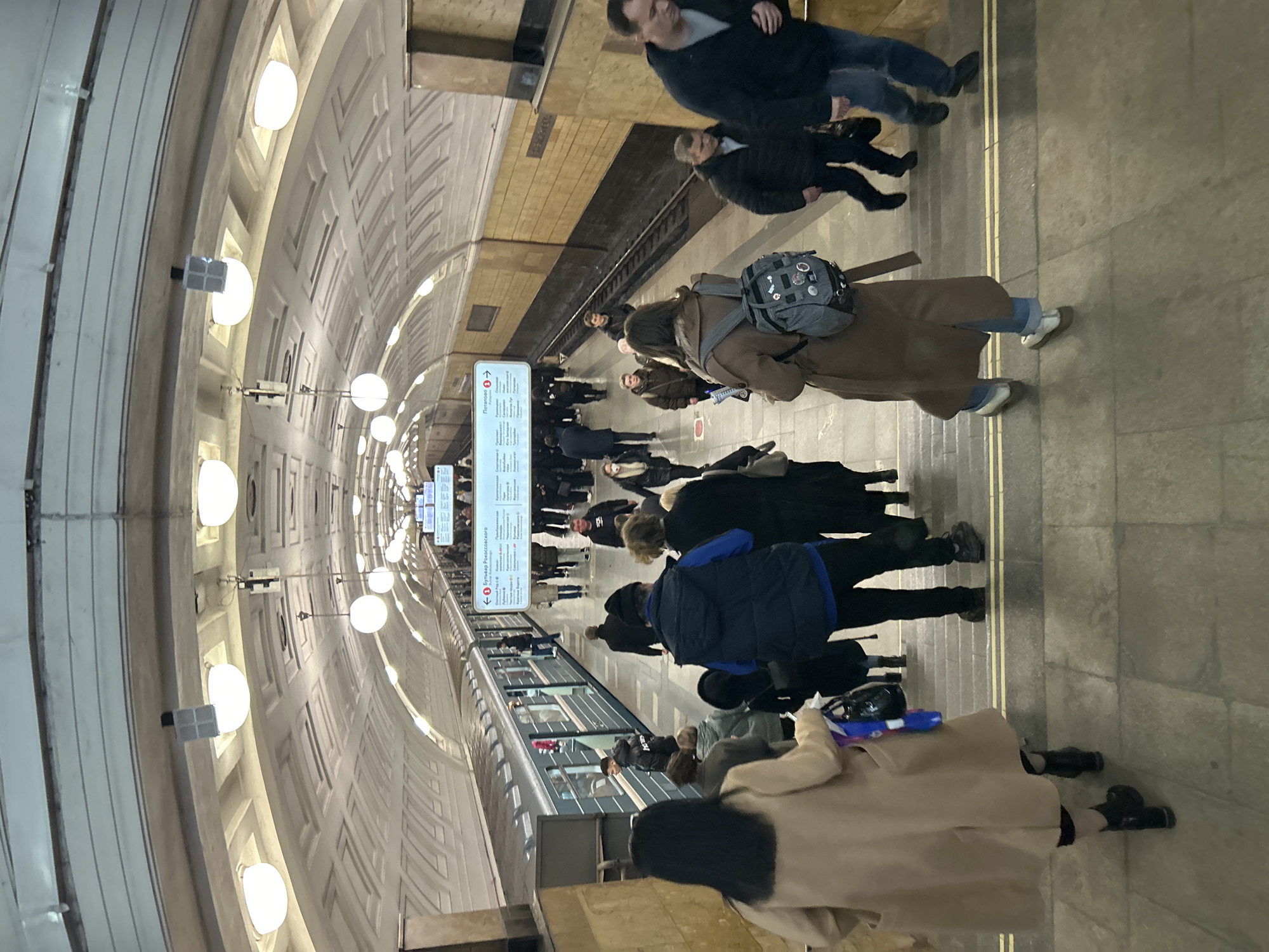 The image captures a crowded Moscow metro station platform. People are waiting for a train on a platform marked by two yellow lines. Signage in Russian indicates station names. The architectural style includes a curved ceiling with evenly spaced globe-shaped lights. Passengers are wearing various types of outerwear, including coats and jackets.