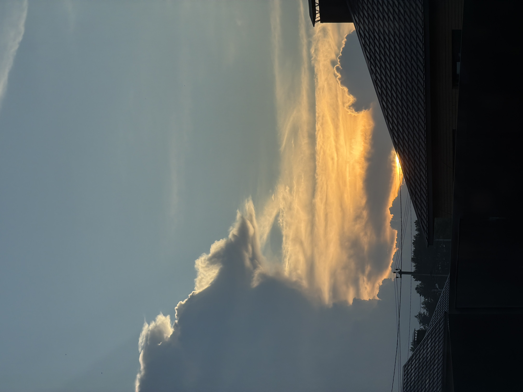 The image depicts a suburban landscape at sunset in Chaikovsky, Russia. The sky is filled with dramatic clouds, with the sun setting behind them, casting an orange glow. In the foreground, silhouetted rooftops of houses and a dark fence are visible. A power pole with wires stretches across the middle ground, with a line of trees behind it. The scene is quiet and serene, capturing the beauty of a natural phenomenon in a residential setting.