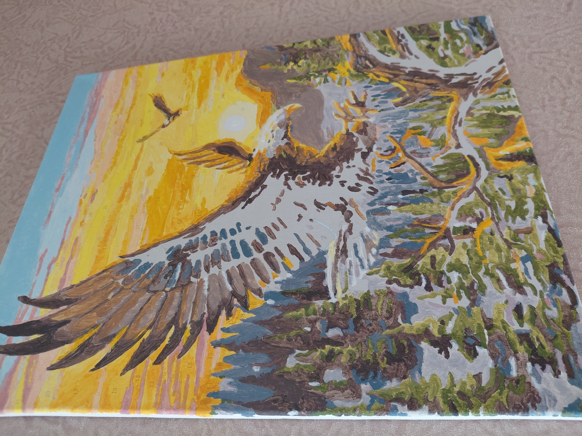 The image is a close-up, angled view of a "paint by numbers" canvas depicting an eagle in flight over a mountainous forest landscape at sunset. The eagle, with outstretched wings, is the central subject, rendered in shades of white, brown, and gray. A smaller eagle is visible in the distance. The background features a vibrant sunset with streaks of orange, yellow, pink, and blue. The landscape below consists of a dense evergreen forest with snow-capped mountains in the background. The canvas is unframed and appears to be leaning against a textured, light-colored wall. There are no visible people, specific landmarks, or any indication of its location in Novosibirsk, Russia.
