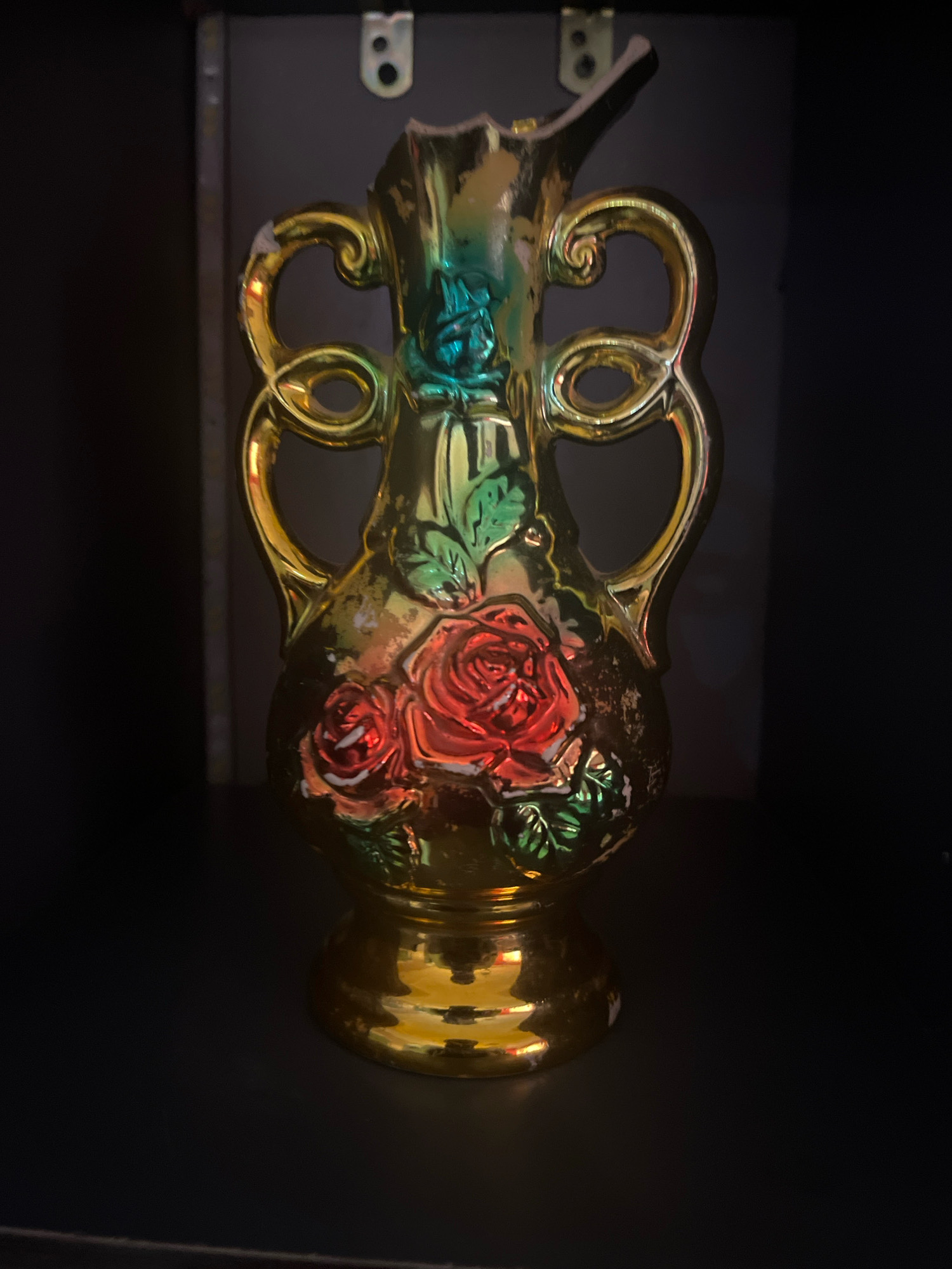 A close-up, slightly low-angle shot captures a gold, iridescent ceramic vase on a dark, reflective surface. The vase is ornate, featuring two handles that curve upwards and outwards, adorned with swirling patterns. A floral motif of red roses with green leaves is embossed on the body of the vase. The top rim of the vase appears to be broken or chipped. The background is dark and indistinct, with a hint of a darker, possibly wooden, structure behind the vase. The lighting casts reflections on the vase and the surface it rests on. The image does not provide any information about its location in Jos, Nigeria.
