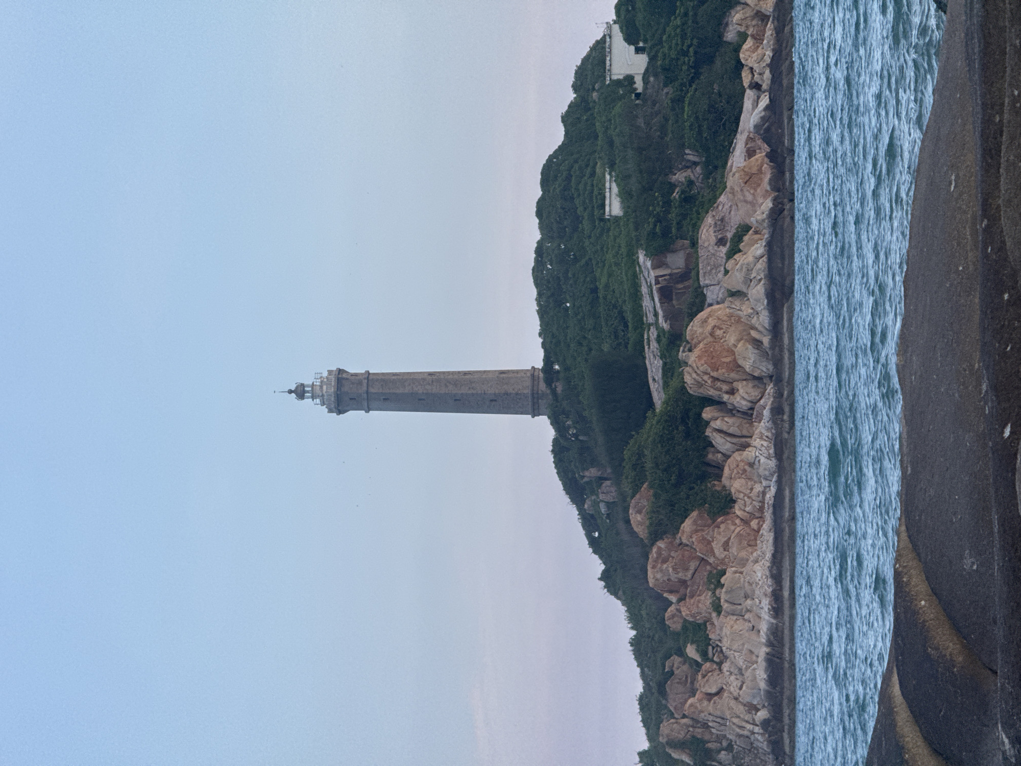 The image depicts a lighthouse situated on a rocky, vegetated island. The lighthouse, constructed of stone, is tall and slender, with a cap at its apex. The island is covered in lush green vegetation and interspersed with large, light-colored rocks. In the foreground, the deep blue water of the ocean is visible, with ripples indicating a gentle movement. The sky above is a pale blue, suggesting it is either early morning or late evening. Two white structures are visible on the island, near the base of the lighthouse, possibly supporting buildings or infrastructure. The overall scene is serene and captures the natural beauty of the coastal landscape.
