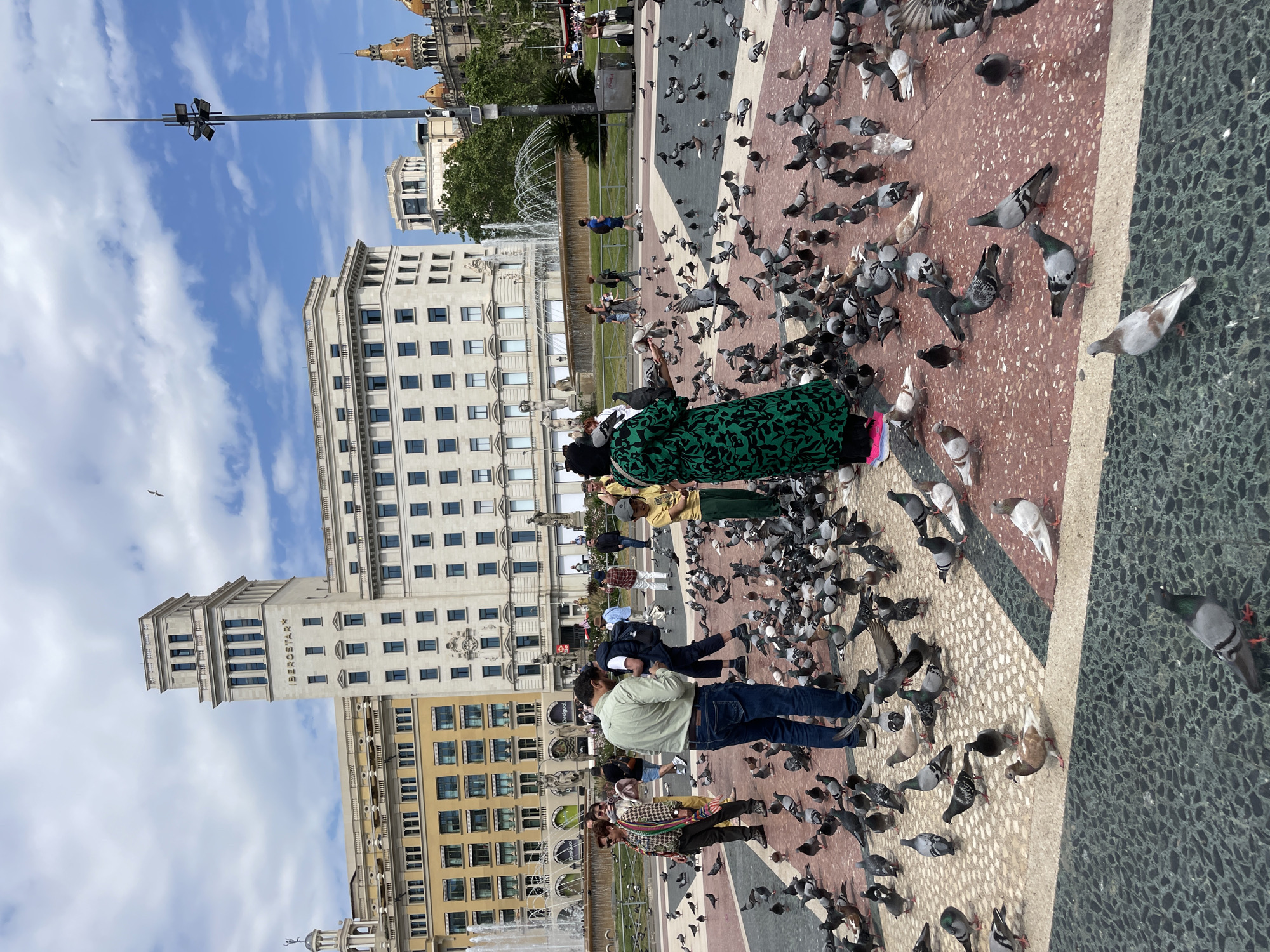 This media file captures a lively daytime scene in a grand urban square, likely in Barcelona, Spain. The foreground features a distinctive patterned ground composed of reddish-brown, light, and dark green/grey tiles, populated by a multitude of pigeons. Several people are present, with a notable number interacting with the birds; one woman in a vibrant green patterned dress stands prominently amidst a large flock of pigeons. Another man in a light green jacket and blue jeans also observes the birds.
In the mid-ground, a large fountain sprays water, adding to the dynamic atmosphere. The background is dominated by impressive classical-style buildings under a bright blue sky dotted with scattered white clouds. A prominent light-colored building with multiple stories and rectangular windows features classical architectural details. Partially visible text on this building appears to read "BERSHKA". Another large building with a yellowish facade is seen further back. In the distance, ornate golden domes and turrets of a grand structure add to the city's iconic skyline. A tall lamppost or street light fixture is visible on the left. The overall scene conveys a bustling yet relaxed urban environment, with people enjoying a sunny day in a historic city square, interacting with local wildlife.