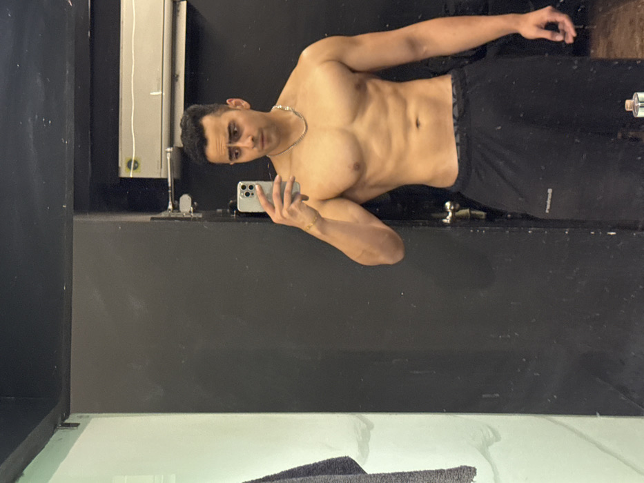 A man is taking a selfie in the mirror. He is shirtless, wearing dark athletic shorts, and a silver chain necklace. The man is tanned with short dark hair. He has a phone in his hand and is looking at his reflection. The background includes a black wall, a white tiled surface, and some plumbing fixtures. The lighting appears to be bright.