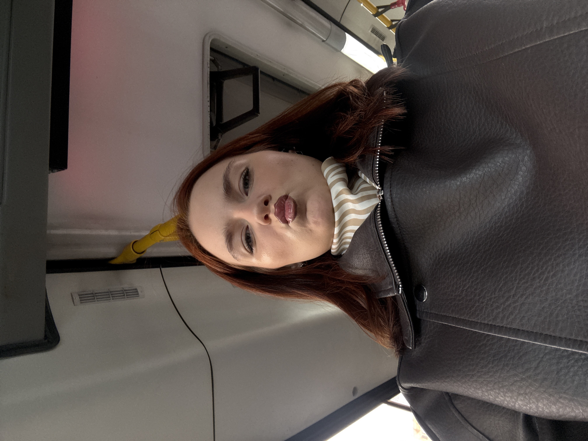 A woman with auburn hair, wearing a brown leather jacket and a beige and white striped scarf, is taking a selfie. She is inside a vehicle, possibly a bus or train, with white interior panels and black accents. There is a yellow handle and a ventilation grill visible on the interior wall. The exact location within Sinyavino, Russia cannot be determined from this image.