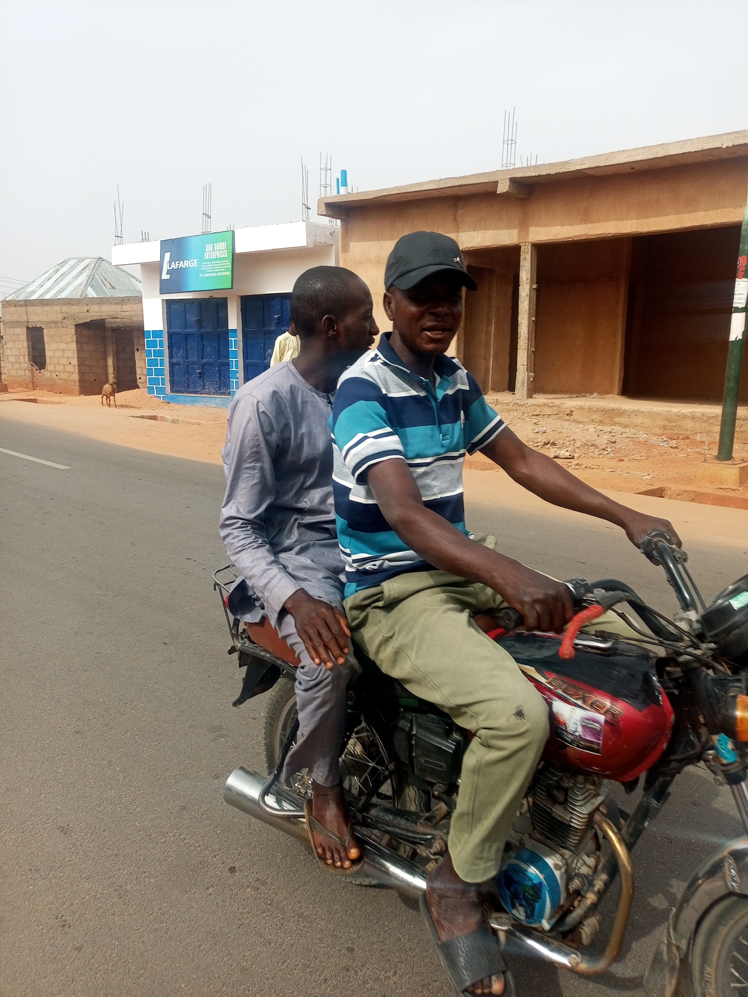 The image captures a street scene in Tempure, Nigeria, featuring two adult males on a motorcycle during bright daylight under a hazy sky. The driver, with dark skin, wears a black baseball cap, a blue and white striped polo shirt, light green pants, and dark sandals. He holds the motorcycle handlebars and appears to be smiling or speaking. Seated behind him is a passenger, also with dark skin, dressed in a light grey traditional long-sleeved tunic and brown flip-flops, looking towards the driver.
The motorcycle features a red fuel tank with a visible "BOXER" label. They are positioned on a paved asphalt road, with a white line visible to the left. In the background, several buildings line the street. A prominent building has a blue and white painted facade with blue double doors. Above these doors, a green sign clearly displays "LAFARGE" and "AAU GOMBE ENTERPRISES," with smaller text below reading "SALES DISTRIBUTION, WHOLESALE & RETAIL." To the left, an unfinished building made of cinder blocks with a corrugated metal roof is visible, and a small brown dog stands in the distance. To the right, another light brown building, possibly residential or commercial, can be seen. The scene depicts a typical daily activity in a semi-urban or rural Nigerian setting.