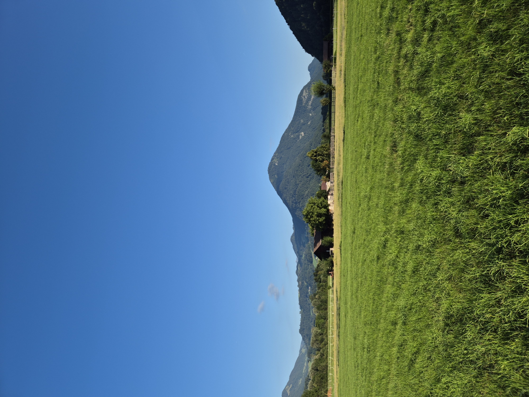 The image depicts a rural landscape under a clear blue sky. In the foreground, a lush green grassy field stretches across the frame. In the middle ground, a wooden fence separates the field from a stretch of drier grass, beyond which are several farm buildings, including a dark-colored barn with a gabled roof. Trees are scattered around the buildings. The background features rolling hills and mountains, some covered in dense forest, with a prominent, rounded peak dominating the center-right of the scene. The scene is tranquil and appears to be a sunny day in Niederndorf, Austria, likely a farm or agricultural area with views of the surrounding natural landscape. There are no people or animals visible in the image.