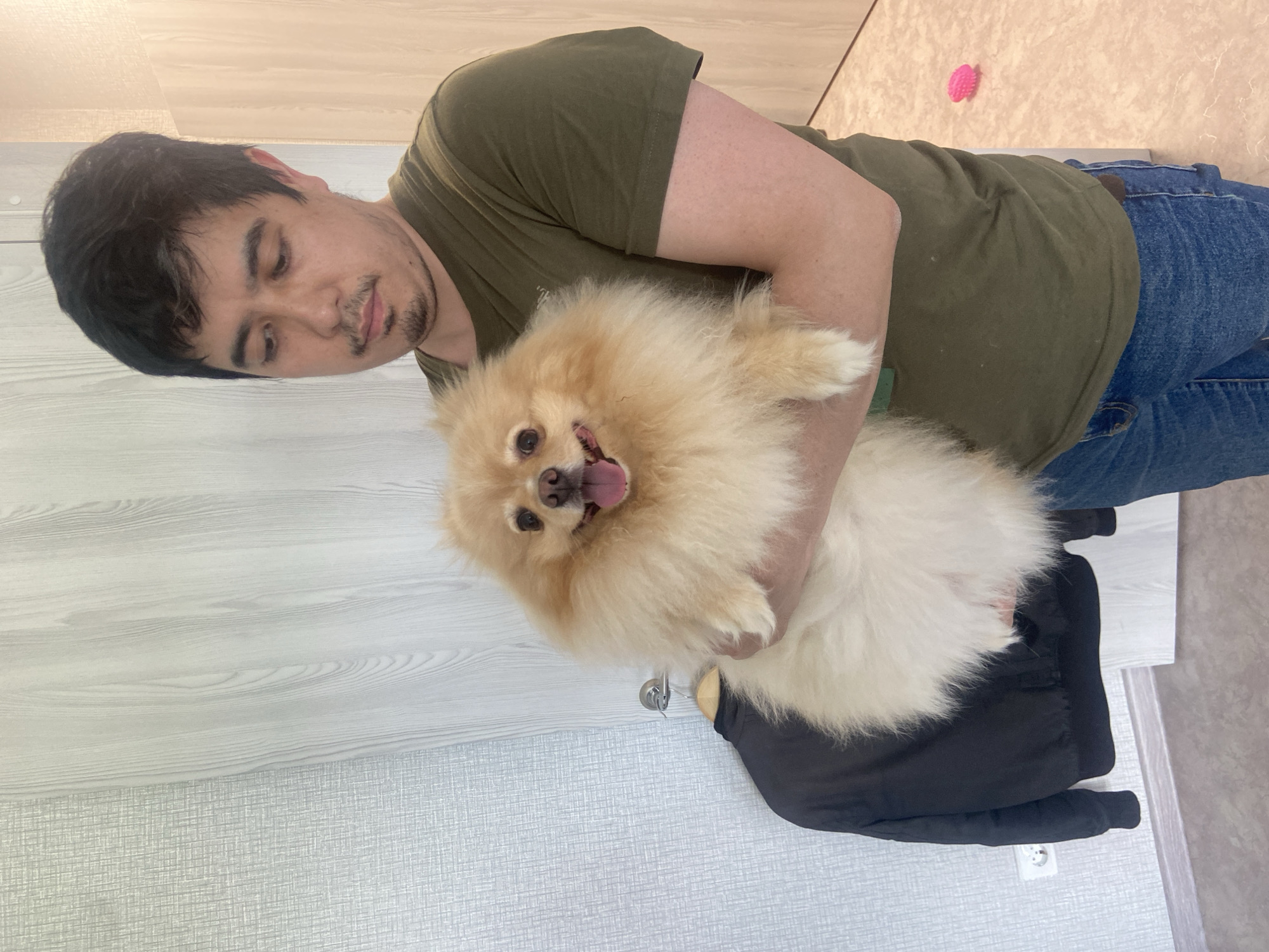 A man holds a fluffy Pomeranian dog in his arms indoors, likely in an apartment in Kazan, Russia. The man is wearing a dark green t-shirt and blue jeans. The dog is light cream colored with its tongue sticking out. Behind them, the wall appears to be decorated with a wood-grain pattern. A black jacket is hanging on a hook on the wall to the left. The floor is a light, neutral color. A small pink object, possibly a dog toy, is visible on the floor in the background.