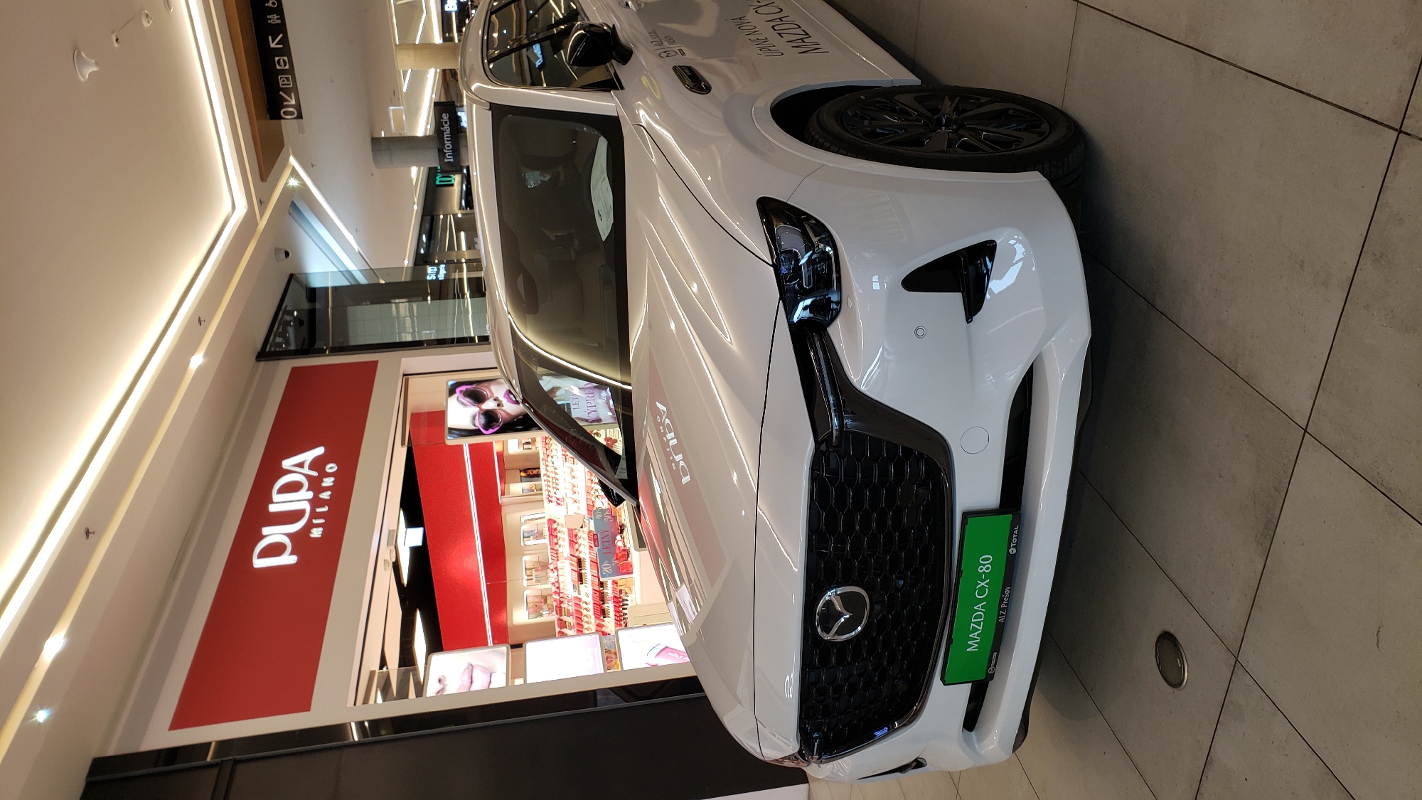 In a brightly lit shopping mall, a white Mazda CX-80 is displayed prominently. The car is parked on a tiled floor, facing slightly to the left, with its front grille, headlights, and badge clearly visible. A green license plate on the front bumper reads "MAZDA CX-80" and also displays "ALZ Prešov" and "TOTAL," indicating its dealership and associated service in Prešov, Slovakia.
Behind the car, a store with a large red sign displays the logo "PUPA MILANO" in white lettering. The store's interior is visible, showcasing rows of makeup products on shelves, with mannequins or displays featuring women's faces. In the background, mall signage includes "Informácie" and pictograms for accessibility and directional information. The ceiling features modern lighting and ventilation systems. The overall environment suggests a retail space within a modern shopping center.