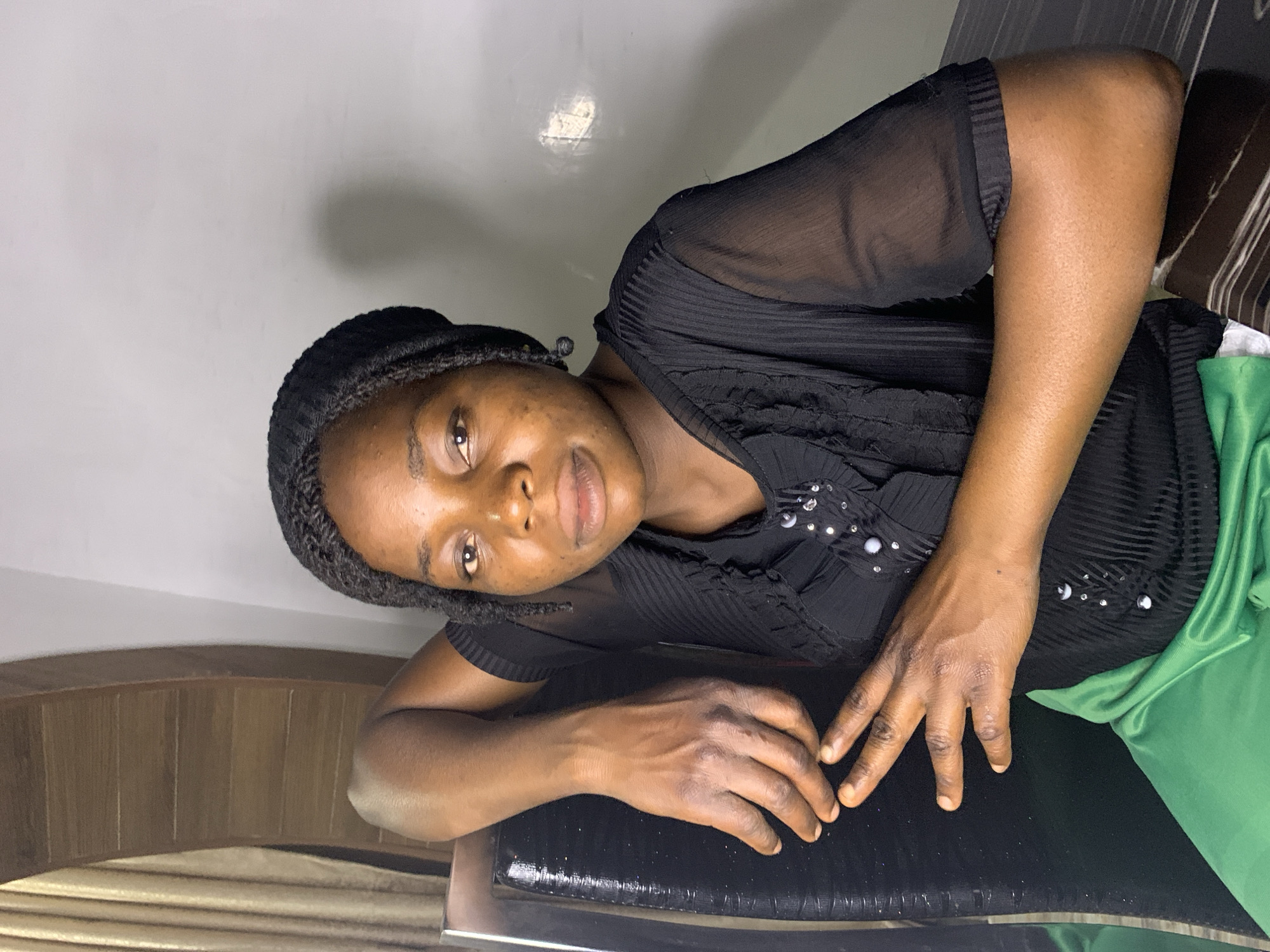 A medium shot captures a Black woman from the chest up, seated. She is wearing a black headscarf and a black, sheer, long-sleeved blouse adorned with sequins and buttons down the front. She is leaning her left arm on the back of a black chair, and her right hand rests on her lap. Her expression is neutral. Behind her, a wooden structure with horizontal slats and beige curtains is visible to the left, and a plain white wall is to the right. The woman is indoors, in a room with modern furnishings. The location is identified as Jos, Nigeria.