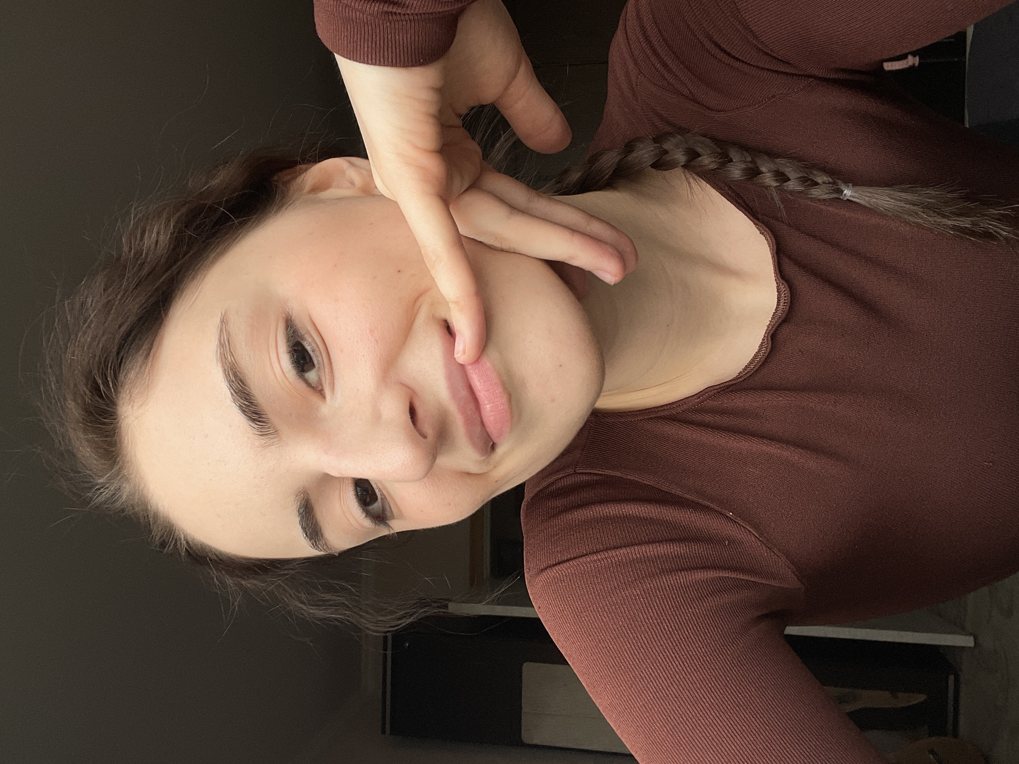 A selfie of a fair-skinned young woman with dark hair, some of which is styled into a braid on her right side. She is wearing a brown, long-sleeved top and is posing with her right hand near her face. The background is dark, and a corner of a white piece of furniture is visible in the lower part of the frame. The location cannot be verified with the image.