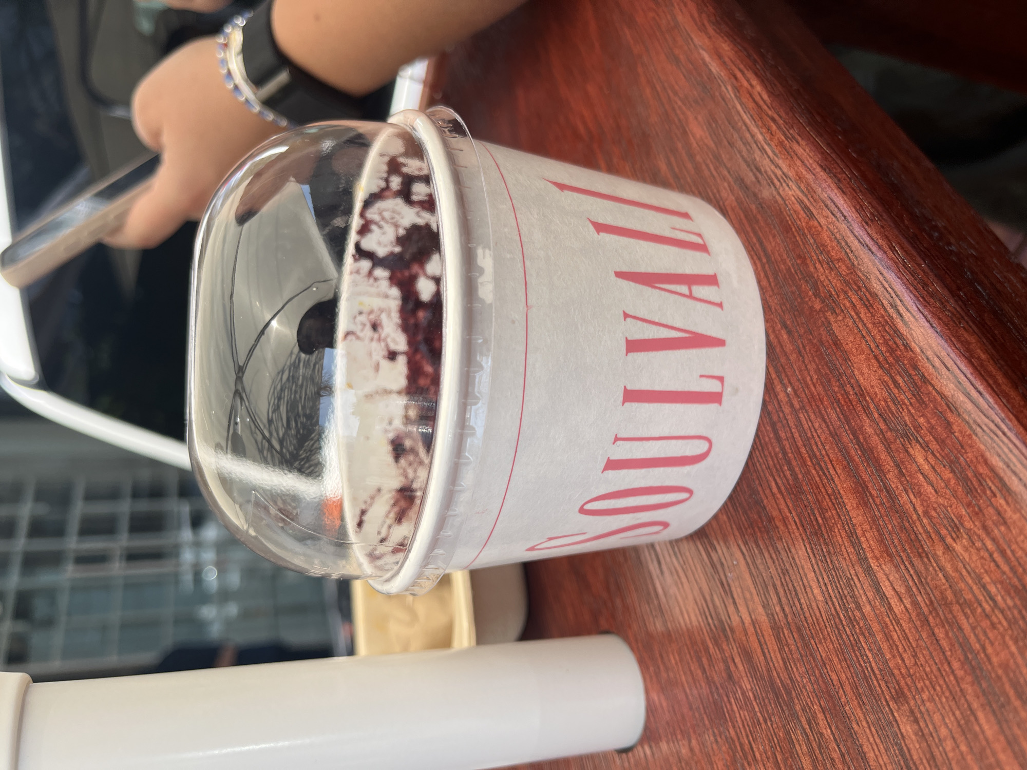 On a wooden table, a clear plastic cup containing a dessert or smoothie with dark-colored ingredients is prominently displayed. The cup has a white paper label printed with pink lettering that reads "SOU VALI" vertically. A transparent, domed lid covers the top of the cup. A person's hand is visible in the background, holding a smartphone. The scene appears to be located in a casual environment, possibly a café or restaurant in the city of Puebla, Mexico. A white table leg is visible at the bottom of the image.