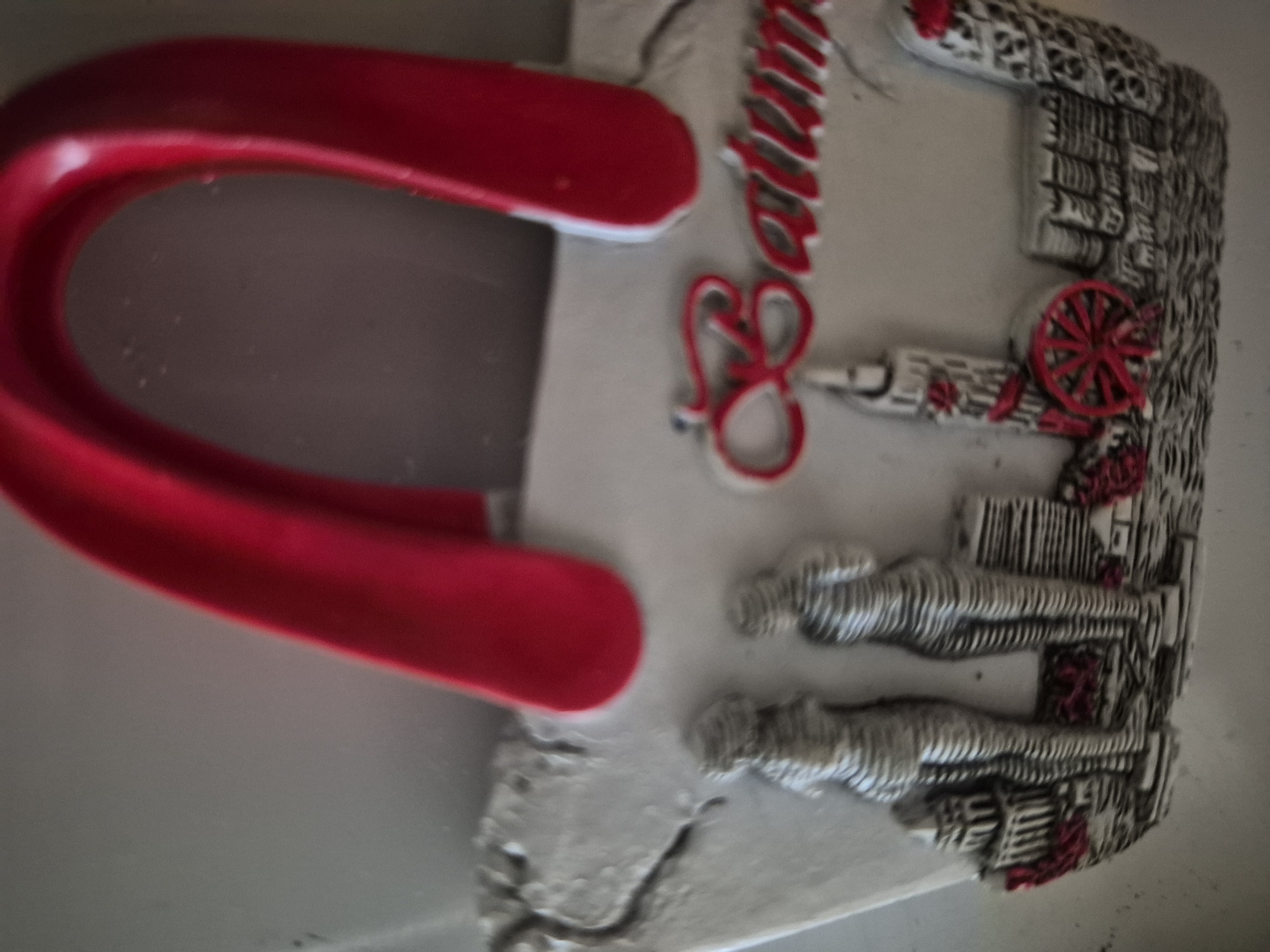 The image shows a close-up of a souvenir, likely a refrigerator magnet, shaped like a white handbag with red handles. The handbag has a decorative design featuring two stylized figures of women standing side-by-side. Below the figures, there is a relief of buildings and architectural elements, including what appears to be a red Ferris wheel. A red cursive inscription across the front of the handbag reads "Batumi". This souvenir is not from Vanadzor, Armenia, but from Batumi, Georgia.