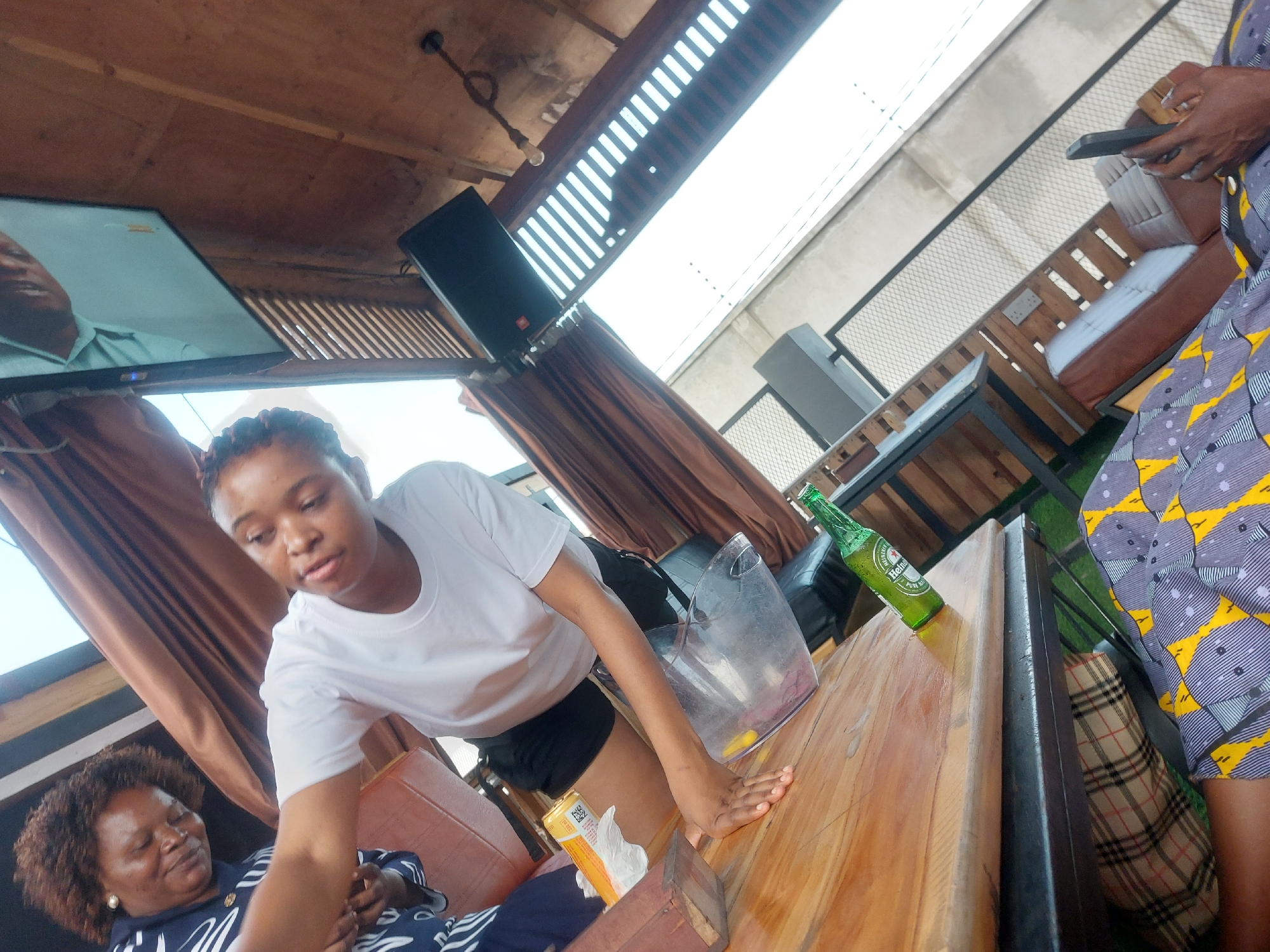 The image shows the interior of a bar or restaurant, likely in Dar es-Salaam, Tanzania, based on the clothing and possible context. A young woman in a white t-shirt and black shorts is reaching towards a wooden table. An older woman in a blue and white patterned top is seated to her left, possibly resting. On the table is an ice bucket, a Heineken beer bottle, and a box of tissues. In the background, there's a television screen, a speaker, and curtains. Another person wearing patterned clothing is partially visible on the right, holding a phone. The seating area has upholstered benches and metal framed tables, possibly in an outdoor or semi-outdoor setting with a slatted roof structure.