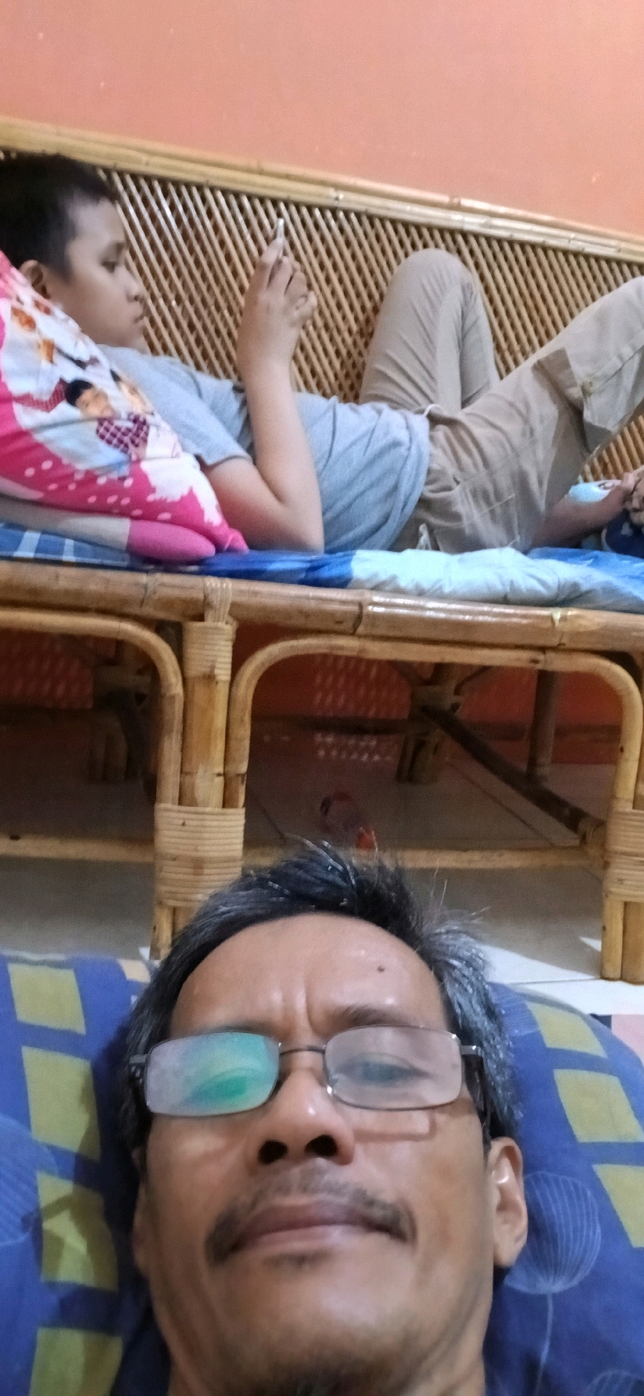 The image is a selfie taken from a low angle, showing the face of an adult male wearing glasses and a mustache. The man is lying down on a blue and yellow patterned pillow. Behind him, a younger person is also lying down on a bamboo-framed daybed, holding a smartphone. The younger person is wearing a gray t-shirt and light-colored pants. A pillow with a pink and white pattern is visible next to them. The background wall is orange. The image appears to be taken indoors, likely in a residential setting in Samarinda, Indonesia. The lighting suggests it is daytime.