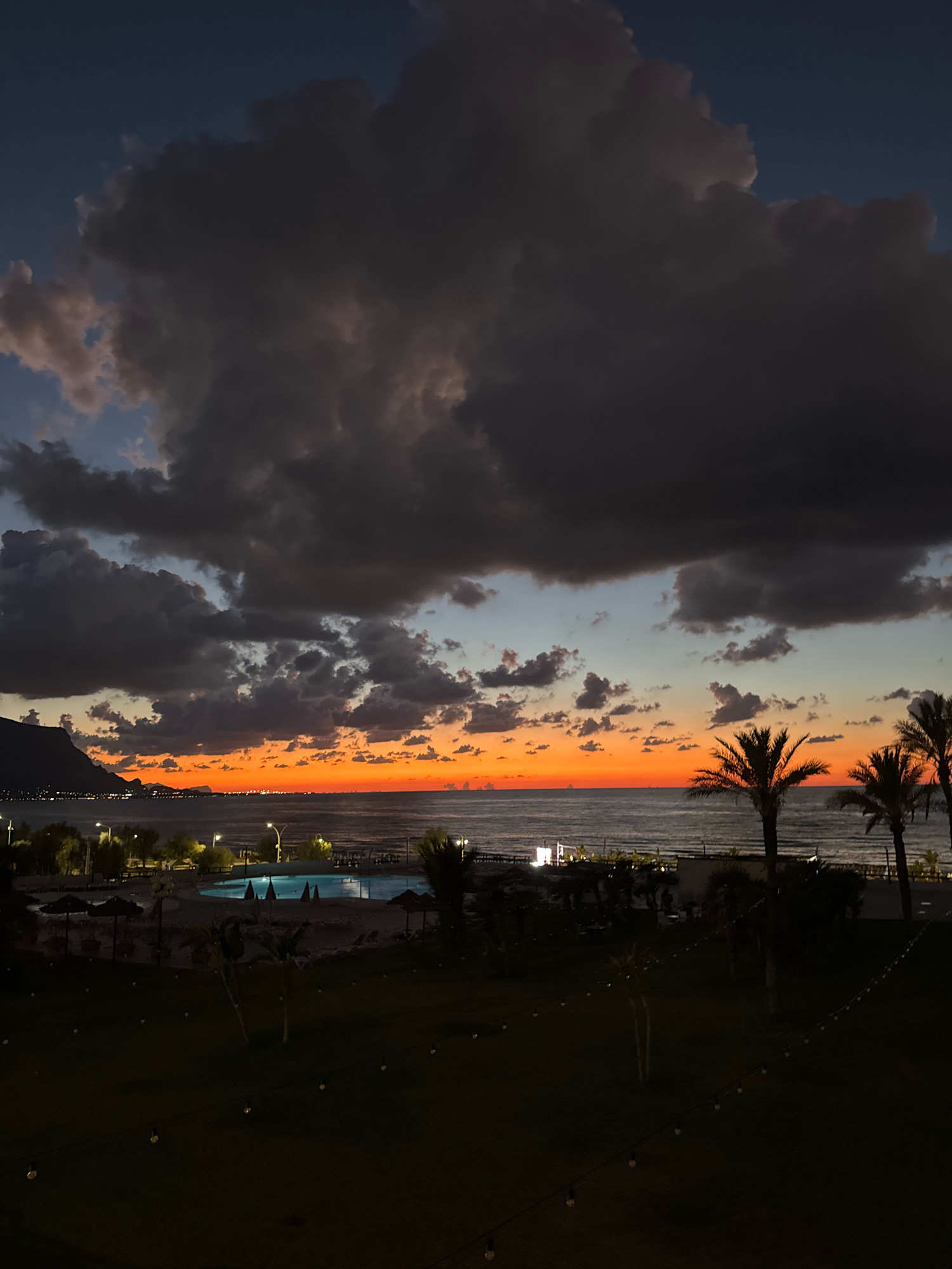 This vertical, high-angle, medium shot captures a dramatic sunset over the coast of Isola delle Femmine, Italy. The sky is a spectacle of color, transitioning from a deep, stormy gray in the upper portion of the frame to a vibrant band of orange and red at the horizon, where the sun has just set. Dark, voluminous clouds dominate the upper sky, creating a sense of depth and intensity.
Below the vibrant sunset, the Tyrrhenian Sea is visible, appearing calm with subtle ripples. The coastline stretches across the mid-ground, illuminated by scattered lights from what appears to be a town or resort. A prominent mountain or hillside juts out from the left side of the frame, silhouetted against the twilight sky.
In the foreground, a landscaped area is visible, mostly in shadow. String lights with visible bulbs are draped across this area, adding a touch of festive ambiance to the scene. A swimming pool, illuminated by underwater lights, is situated in the mid-ground, with lounge chairs and umbrellas scattered around its perimeter. Palm trees are interspersed throughout the scene, their silhouettes dark against the colorful sky. The overall impression is one of a peaceful evening at a coastal resort, with the natural beauty of the sunset creating a striking backdrop. There are no people visible in the frame.
