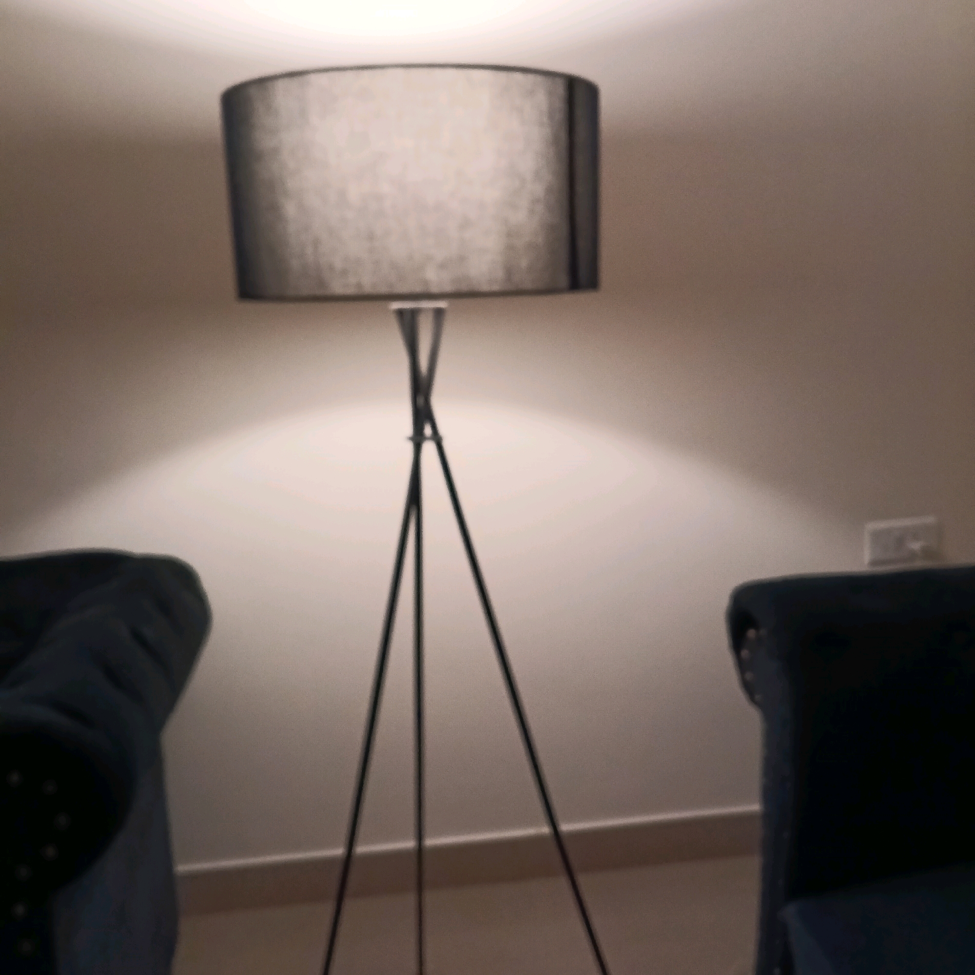 The image shows a dimly lit interior scene, likely a room. In the center, a tall, black tripod floor lamp with a dark grey cylindrical lampshade is illuminated, casting a warm glow on the wall behind it. The wall is a neutral, light color, and a white electrical outlet is visible on the right side. On either side of the lamp, the dark blue upholstery of two chairs is partially visible. The chair on the left is closer to the viewer and its armrest, detailed with small studs, is prominent. The chair on the right is mostly out of frame. The floor appears to be a light color, with a light-colored baseboard running along the wall. No people are visible in the image. The scene does not contain any recognizable landmarks or features that would definitively place it in Bengaluru, India.