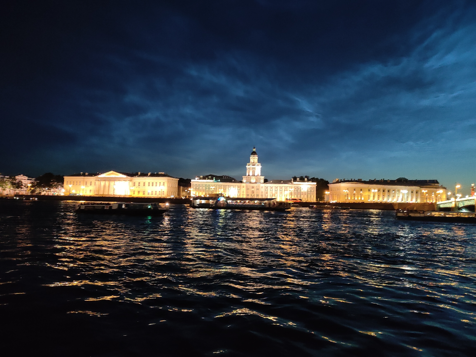 This is a nighttime long-shot photograph taken from a body of water, likely a river or canal, in Saint Petersburg, Russia. The scene features a row of illuminated neoclassical buildings lining the opposite bank, with a prominent bell tower rising above the central structure. The water in the foreground reflects the lights of the buildings and the sky, creating a textured, rippling surface. The sky above is a deep, dark blue, with wispy, illuminated clouds suggesting either twilight or artificial lighting interacting with atmospheric conditions. A few boats are visible on the water near the shore, and a bridge can be seen in the far right of the frame, also illuminated. The overall impression is one of a serene, majestic urban landscape at night.