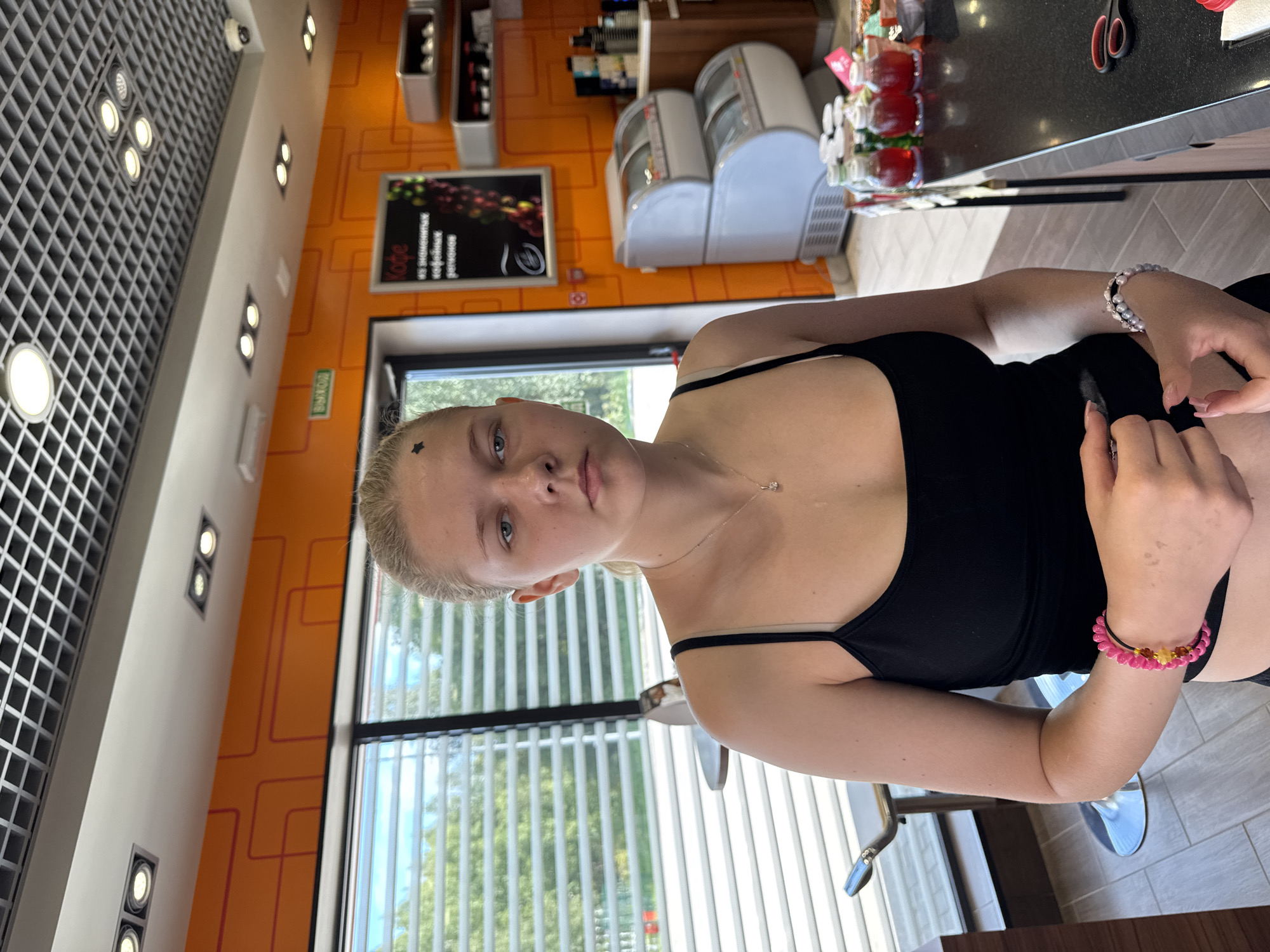 A young Caucasian female with blonde hair pulled back into a ponytail and a small black star sticker on her forehead stands in what appears to be a cafe or convenience store. She is wearing a black tank top with thin straps and a delicate silver necklace with a single pendant. Her right arm is bent, and her hands are clasped in front of her, with bracelets on both wrists – a pink and yellow beaded one on her right and a pearl-like one on her left. The background features an orange wall with a graphic design, a window with blinds, a black counter, and various items on display, including bottles of red liquid, two stacked refrigerators, and a sign with text that appears to be in Russian. The ceiling has a grid pattern with embedded lights. The overall lighting is bright, suggesting daylight. The location is identified as Marchugi, Russia.