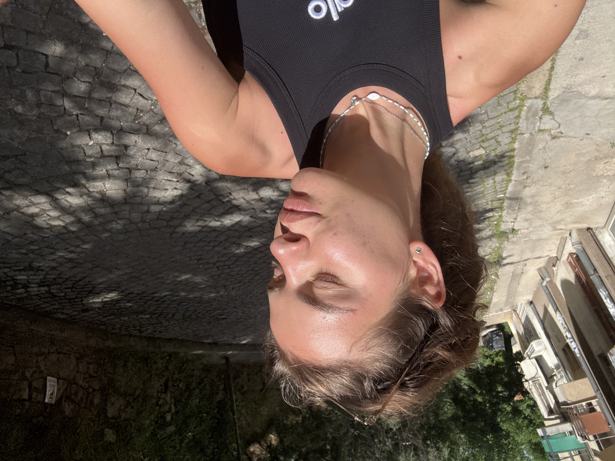 A medium, full shot, rotated, shows a light-skinned person wearing a black top with the word "Alo" in white lettering, a white and turquoise beaded necklace, and a silver necklace. The person is upside down with their eyes closed. The background is a cobblestone street with shadows from trees, buildings, and foliage. A section of a building with windows is visible. The location appears to be outdoors.