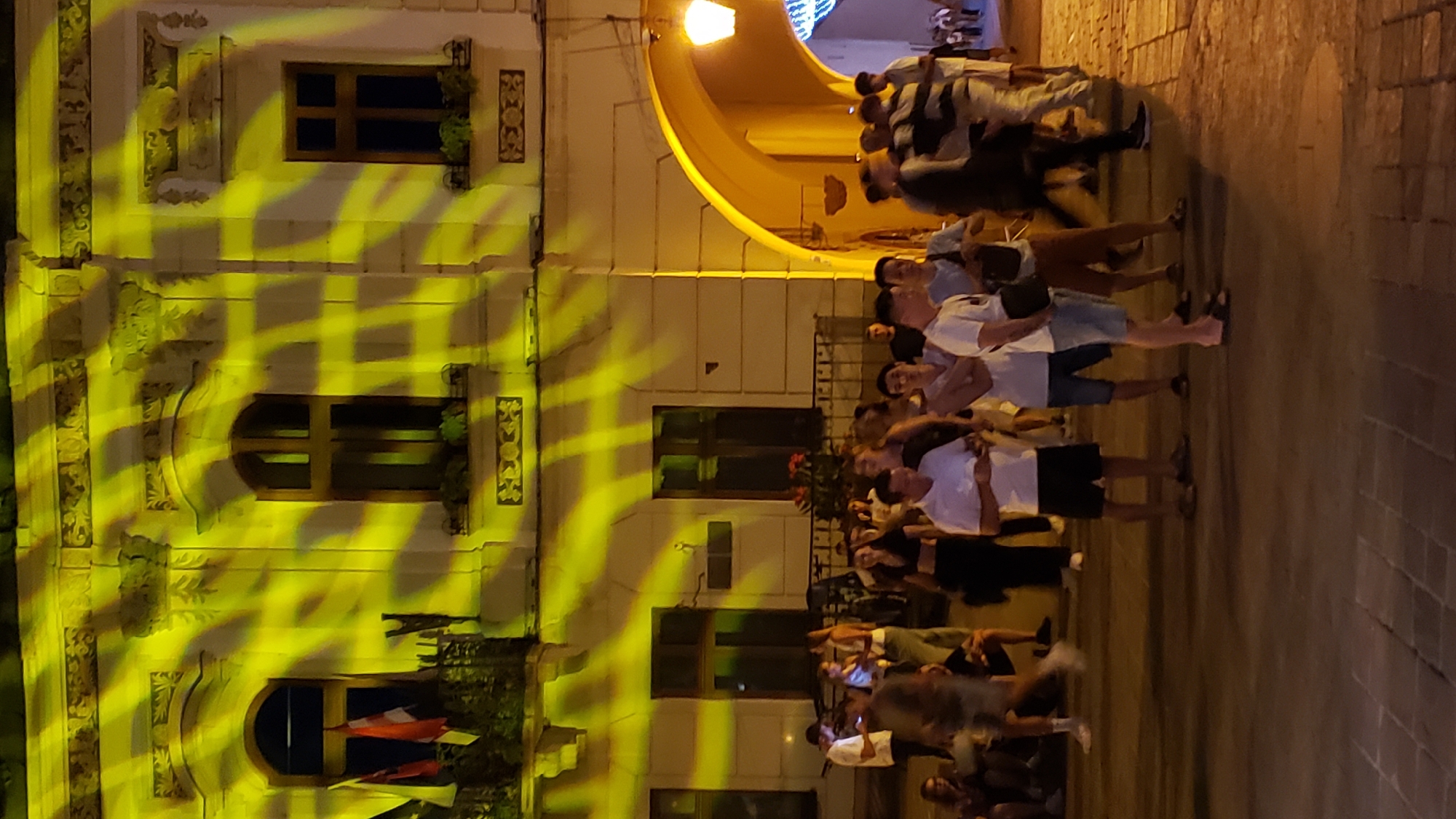 The image depicts a nighttime scene in front of a building adorned with yellow, patterned lighting effects. A group of people are gathered on a cobblestone street. The building features arched entrances and windows, with flags visible on a balcony. The overall atmosphere suggests an outdoor event or gathering. The scene is located on Hlavná Street in Prešov, Slovakia.