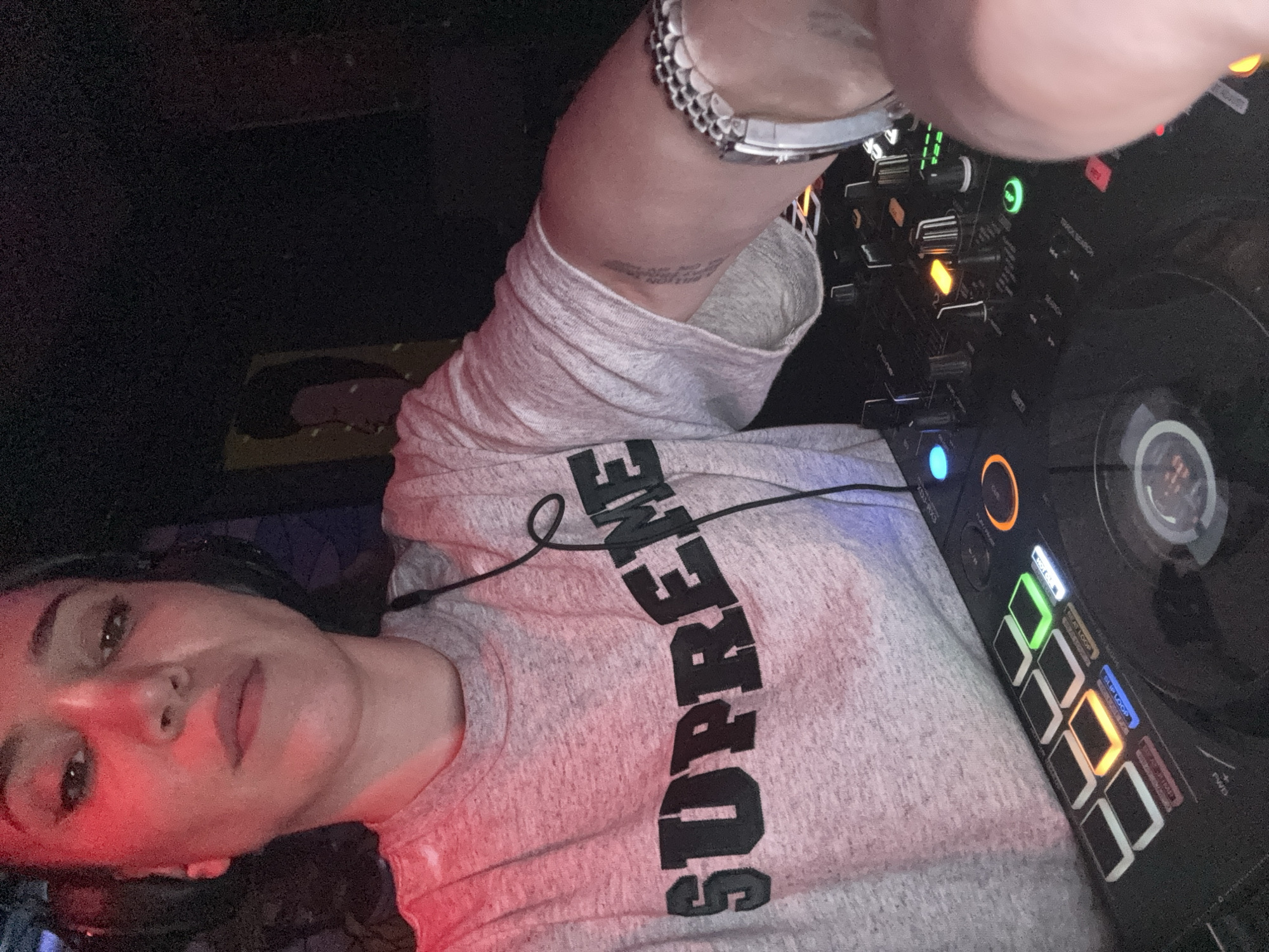 This image captures a person, likely female, engaged with DJ equipment in a dimly lit indoor venue, specified as being in the city of Moscow, Russia. The individual is positioned from the chest up, facing the camera for a selfie-style shot. She wears a grey marl t-shirt featuring the word "SUPREME" in dark block letters across the chest. Headphones are worn over her ears, with a black cord visible descending over her shirt. Red ambient lighting illuminates her face and upper torso, while her expression is neutral. A silver-toned watch with a metal link band is visible on her right wrist, and a vertical tattoo is present on her inner left bicep. Her right hand is actively positioned over a black DJ controller, interacting with its illuminated buttons and knobs. Visible elements on the controller include square performance pads glowing orange, green, and white, alongside other blue, orange, and green indicator lights and a partially visible jog wheel with a central display. The background is dark and indistinct, with a faint, abstract artwork or poster depicting a profile visible to the upper left. The environment strongly suggests a nightclub or similar entertainment establishment.