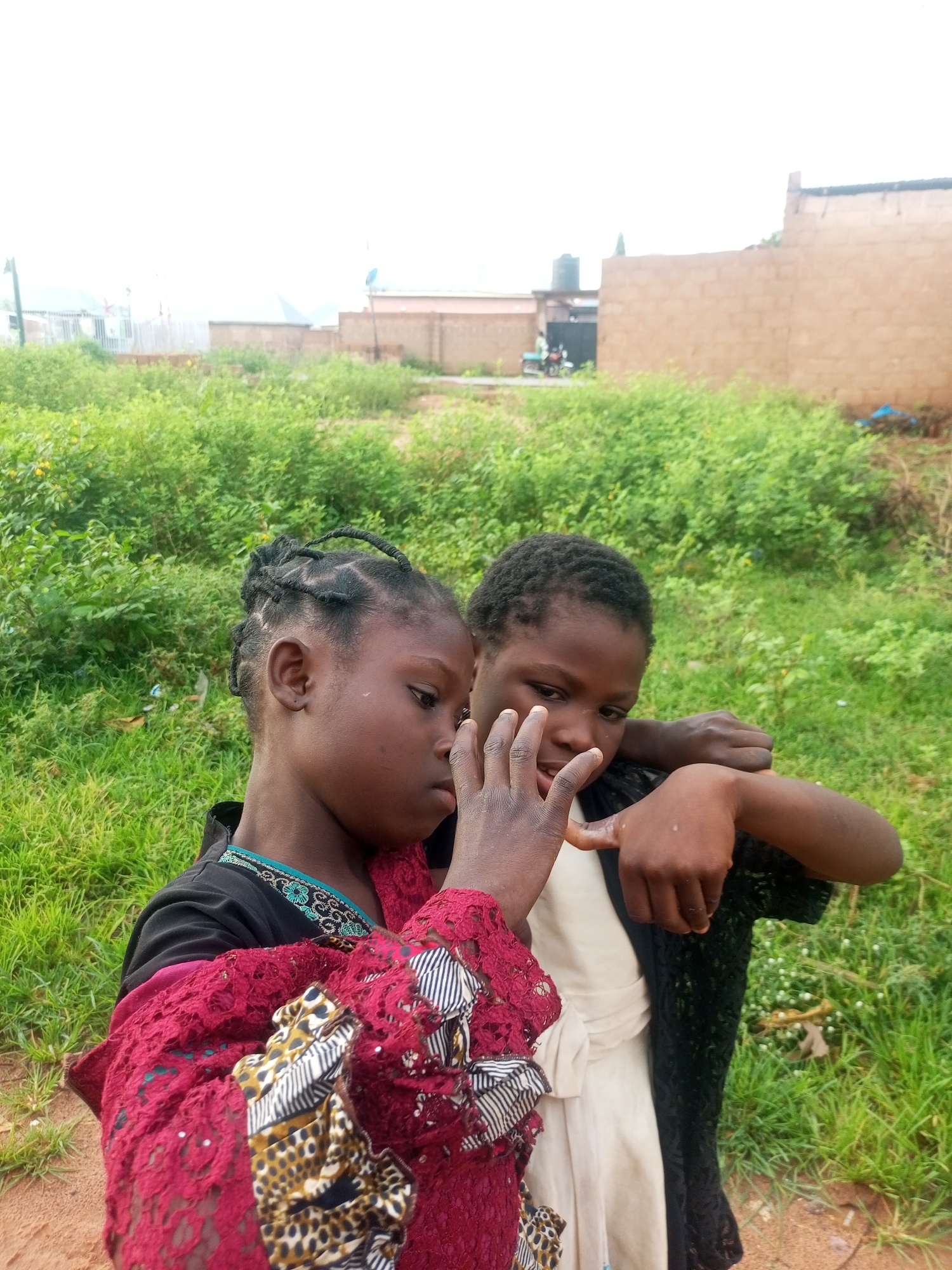 The image depicts two young girls in an outdoor setting in what appears to be a rural or semi-urban area within Tempure, Nigeria. The girls are standing close together, with the girl on the left, wearing a red and patterned garment over a black top, leaning towards the girl on the right. The girl on the right wears a light-colored garment and a black lace overgarment. The girl on the left has her hair styled in braids, and both girls are looking in the same direction. Their hands are positioned near each other, suggesting an interaction. The background consists of lush green vegetation, including bushes and grass, with hints of buildings and a water tank visible in the distance. The overall lighting suggests an overcast day.