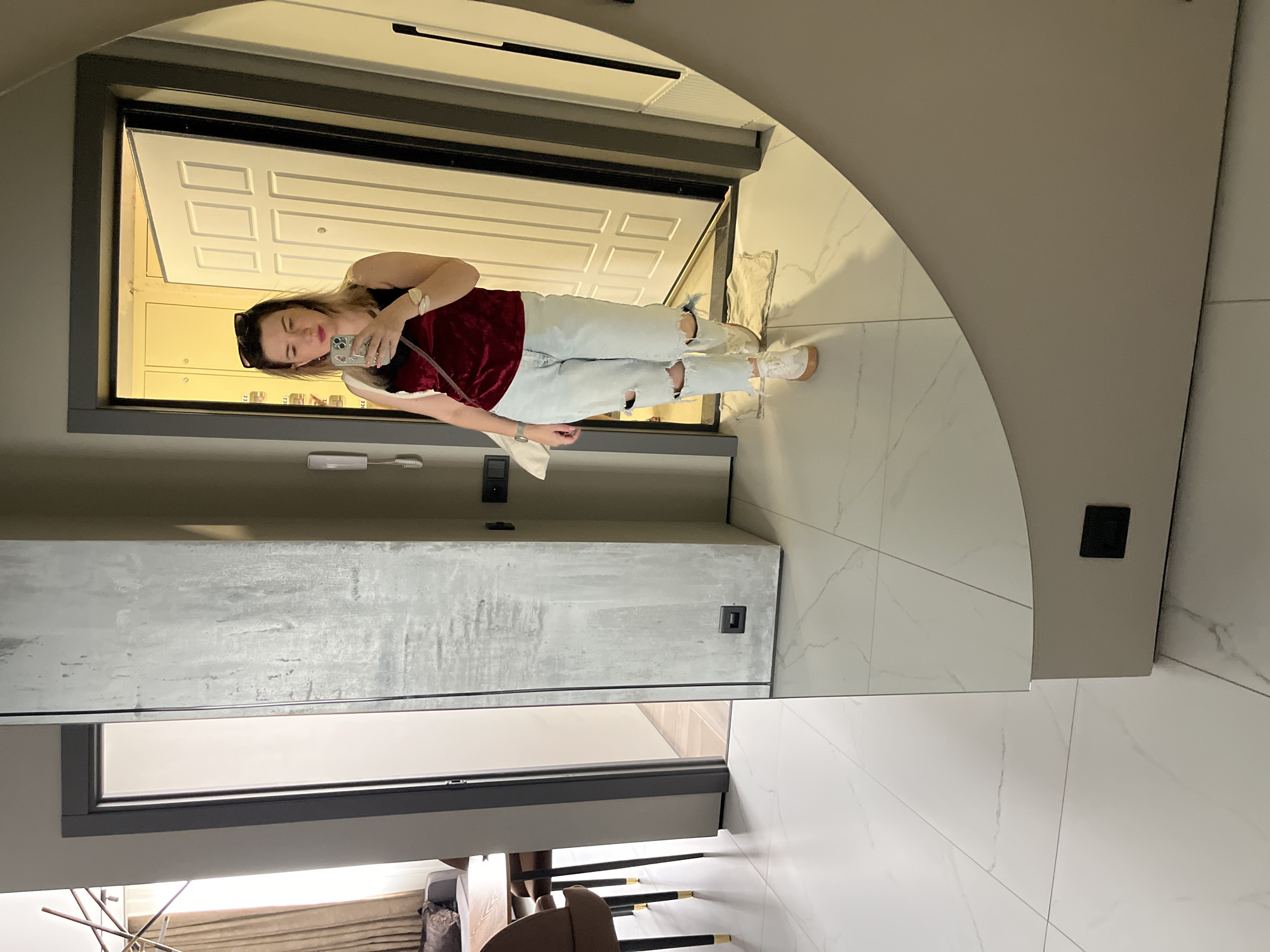 A woman stands in a hallway, taking a selfie in a large, round mirror. She is wearing a dark red velvet tank top, light blue ripped jeans, and white sneakers. She has dark hair and is wearing sunglasses on her head. A white handbag hangs from her left shoulder. The hallway features a gray textured wall on the left and a white door with paneling on the right. The floor is tiled in a marble pattern. In the background, to the left of the woman, is a glimpse of a living or dining area with a sofa, a table, and chairs. The scene suggests an interior setting, likely a residential apartment or a modern hotel lobby. The image is not explicitly tied to Sochi, Russia, but the context suggests a contemporary urban environment.