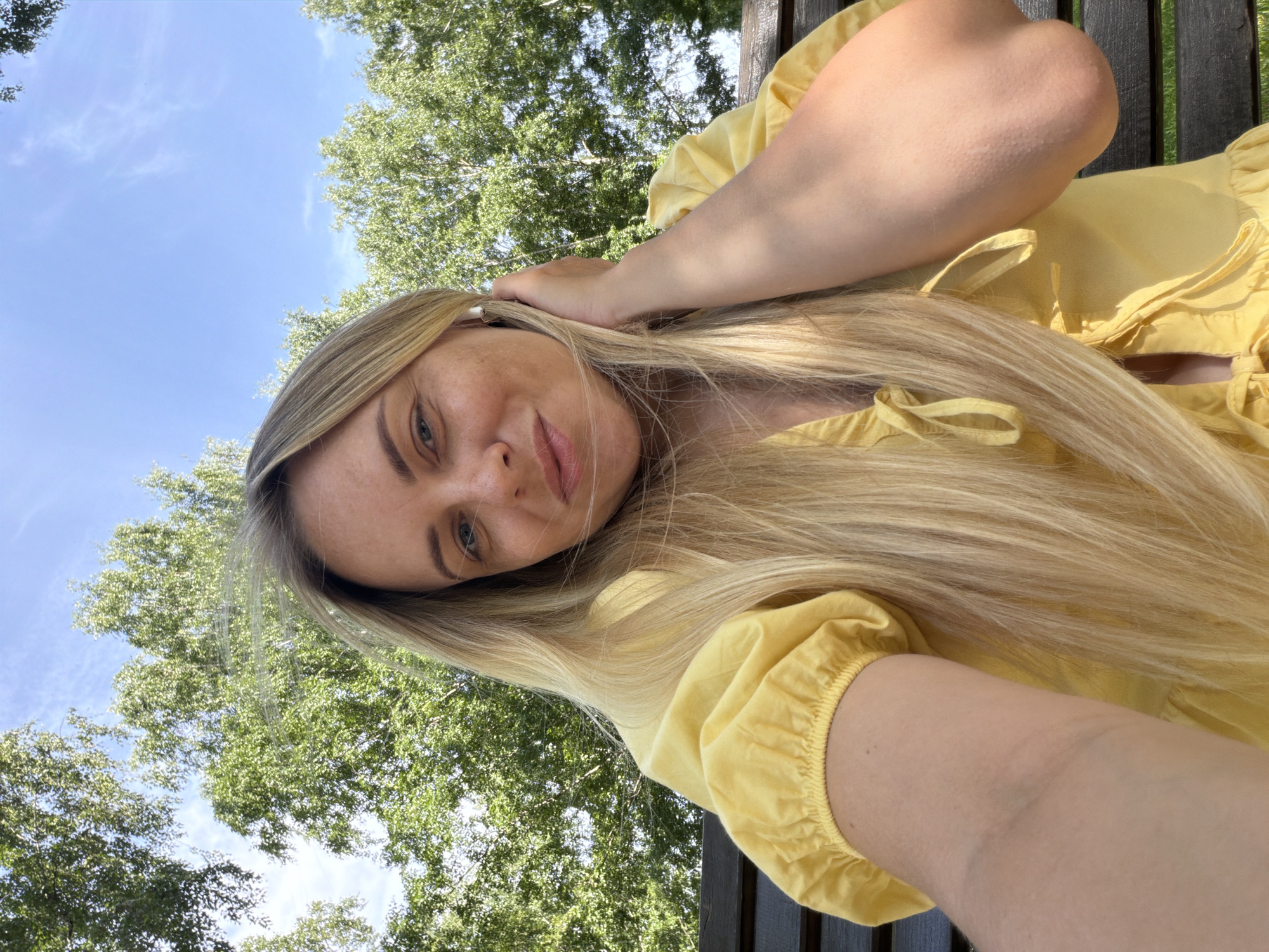 A woman with long blonde hair and fair skin is captured in a selfie. She is wearing a yellow dress with puffy sleeves and a V-neckline. Her right hand is raised to her ear, touching her hair. She is sitting on a dark wooden bench, with lush green trees and a bright blue sky in the background. The lighting suggests it is daytime, likely outdoors. The scene appears to be in a park or a natural setting. There is no visual information to indicate the location in Votkinsk, Russia.