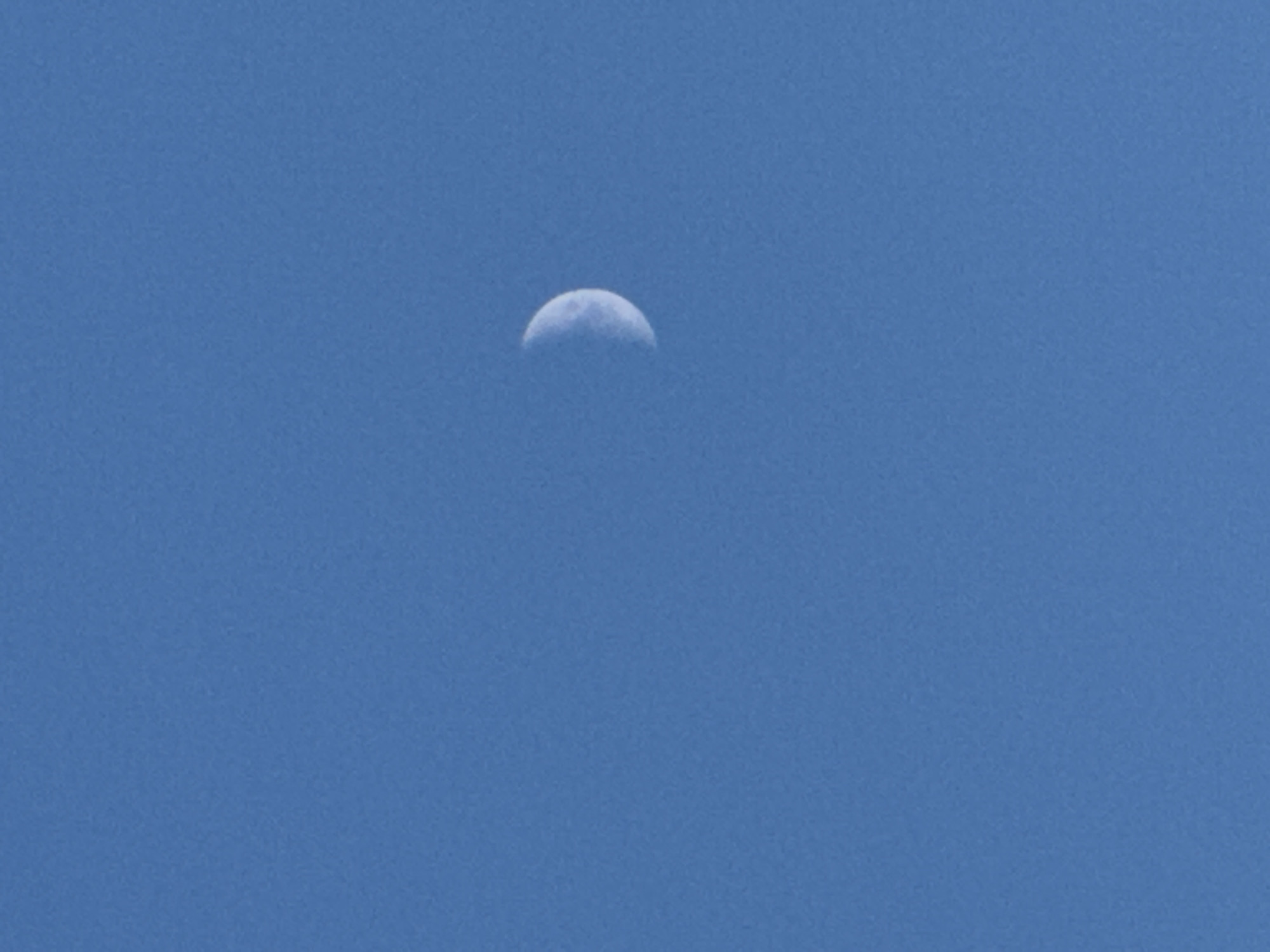 The image shows a clear blue sky with a crescent moon visible in the upper right quadrant. The moon is partially illuminated, showing a curved, bright edge and a darker, shadowed portion. The sky is uniform in color and texture, suggesting it was taken during the daytime or twilight. No other celestial bodies or atmospheric phenomena are apparent. The image does not contain any information about the city of Прикамский, Russia, or any of its features.
